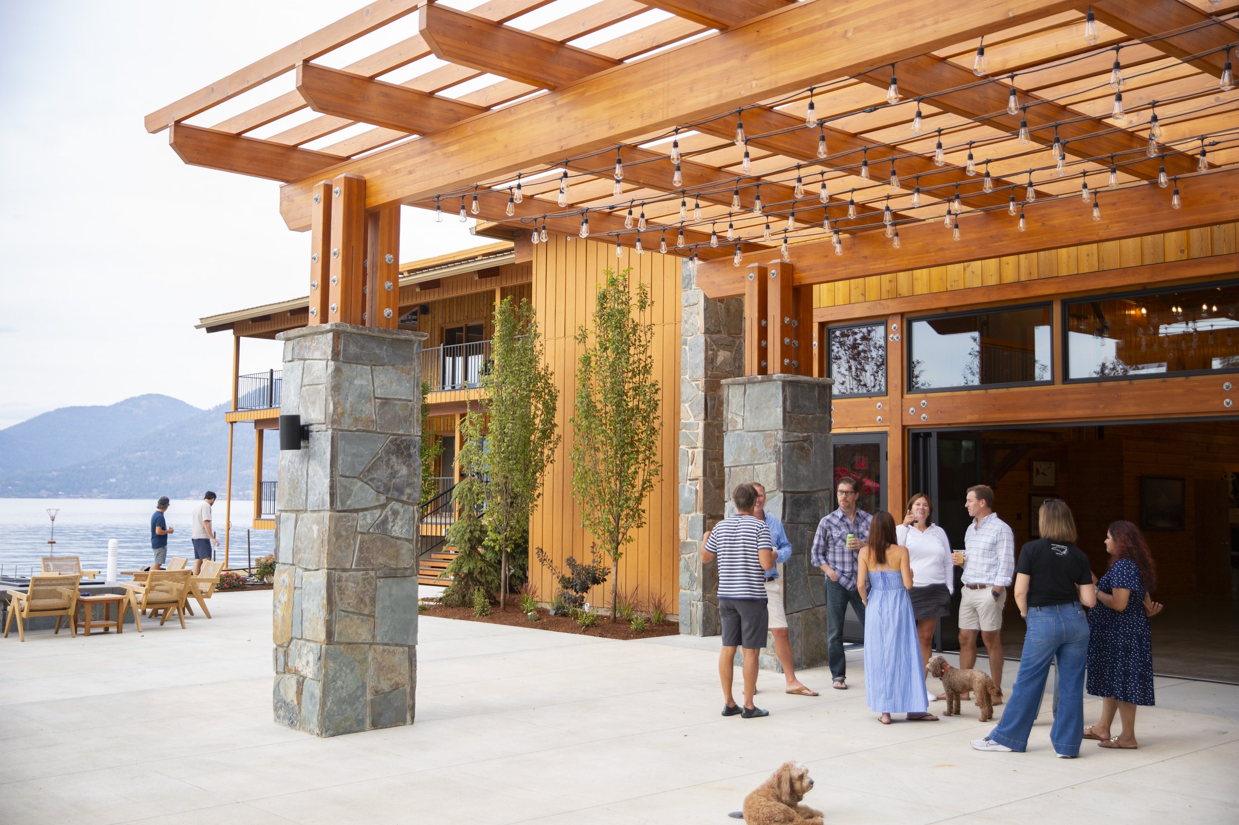 People gathering outside a building with wooden beams and string lights, overlooking a body of water in the background.