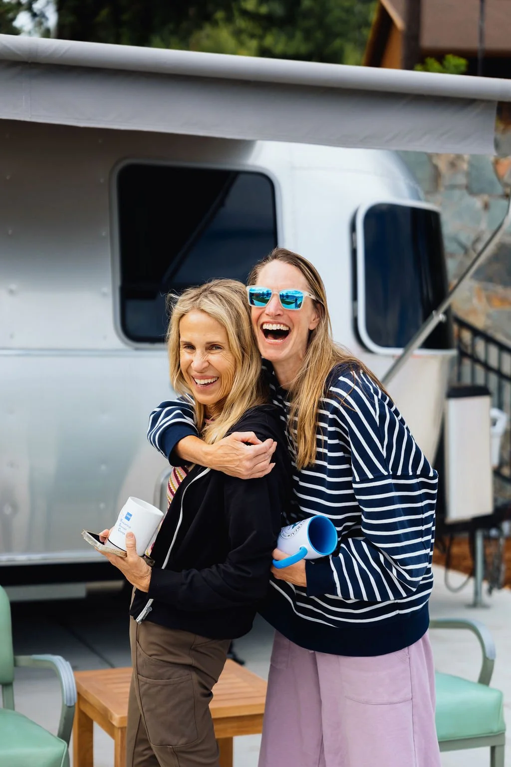 Two women smiling and embracing each other outside near a trailer, holding cups, with chairs and a small table nearby.