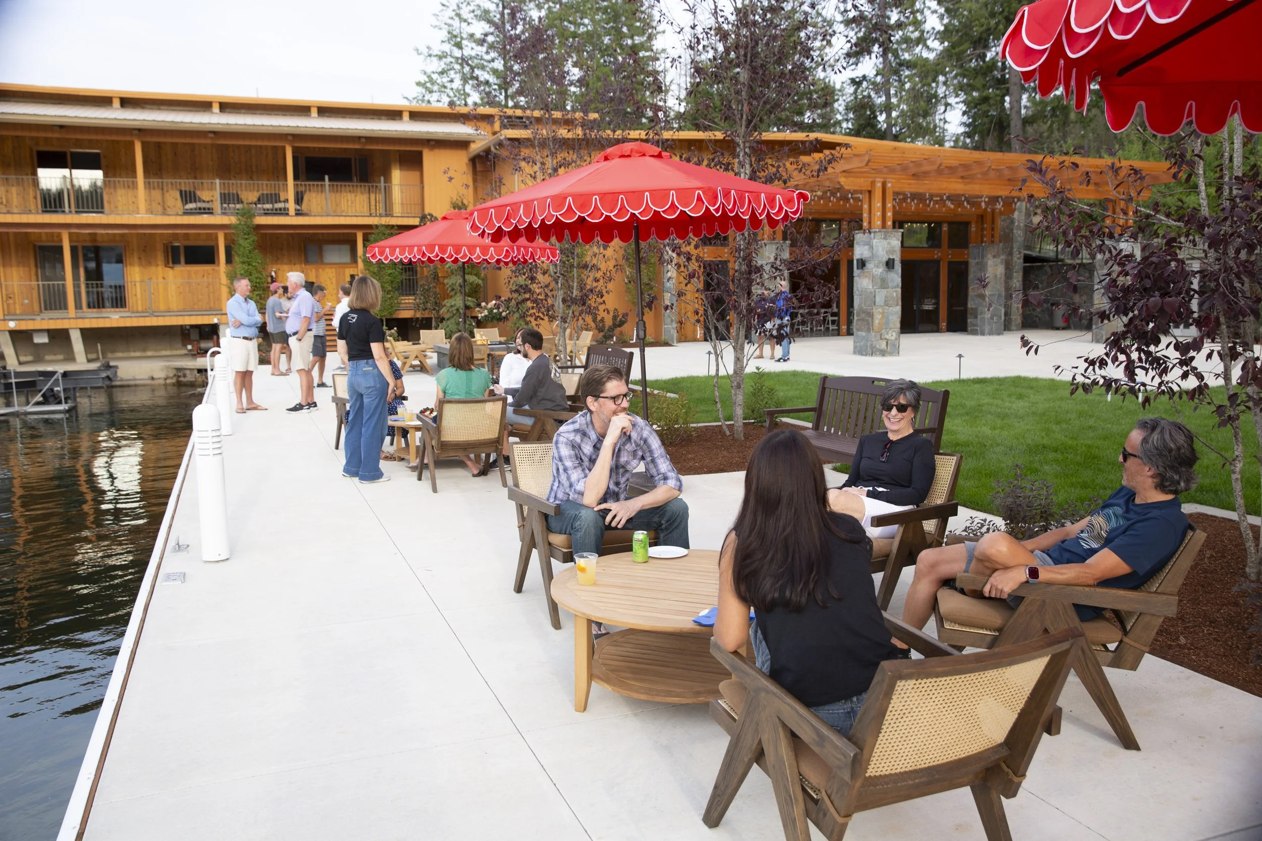 People enjoying outdoor gathering with tables, chairs, and large red umbrellas near a waterfront building with a balcony, trees, and a dock.
