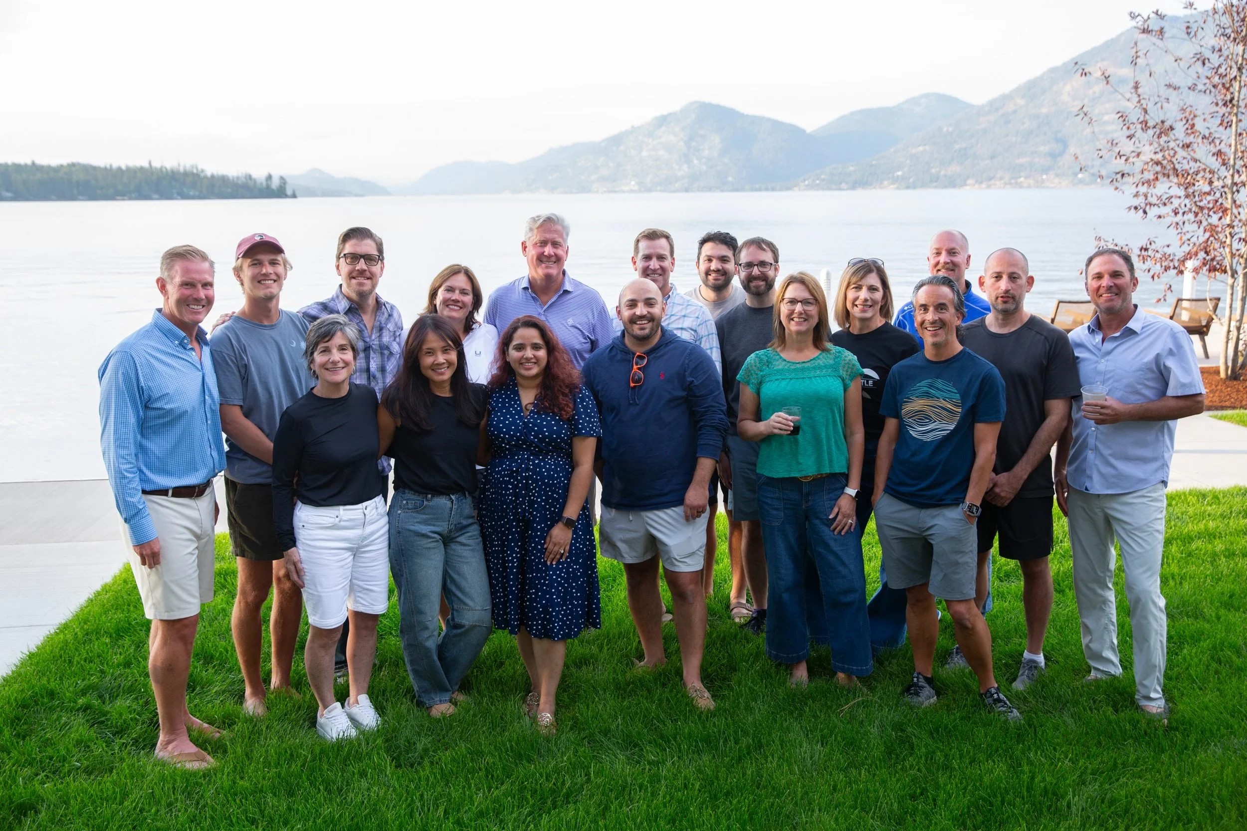 A group of twenty diverse people smiling, standing outdoors on a grassy area near a lake with mountains in the background, during daytime.