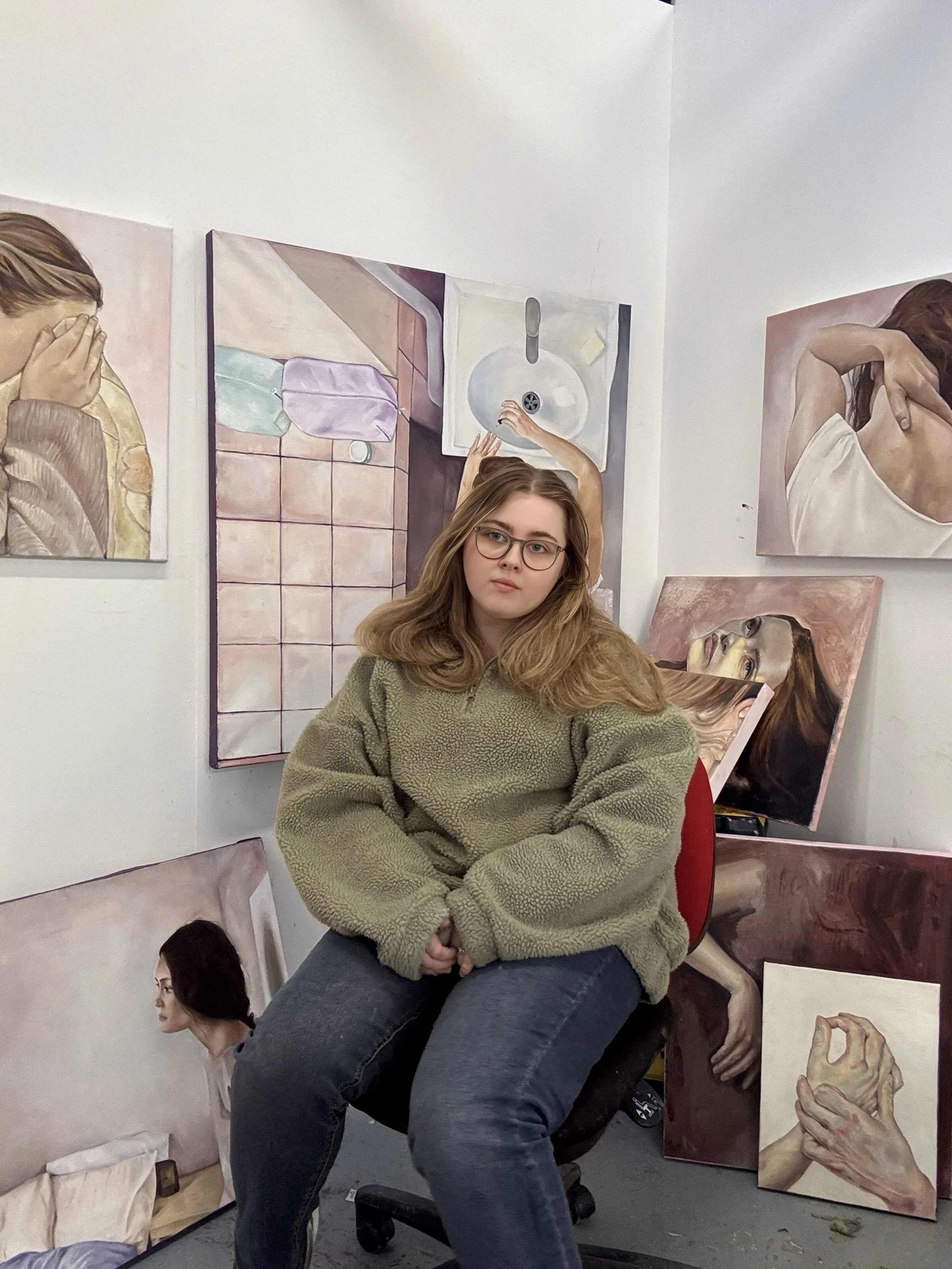 A young woman with glasses and long hair, wearing a beige sweater, sitting in front of a white wall decorated with multiple paintings of women and hands, in an art studio.