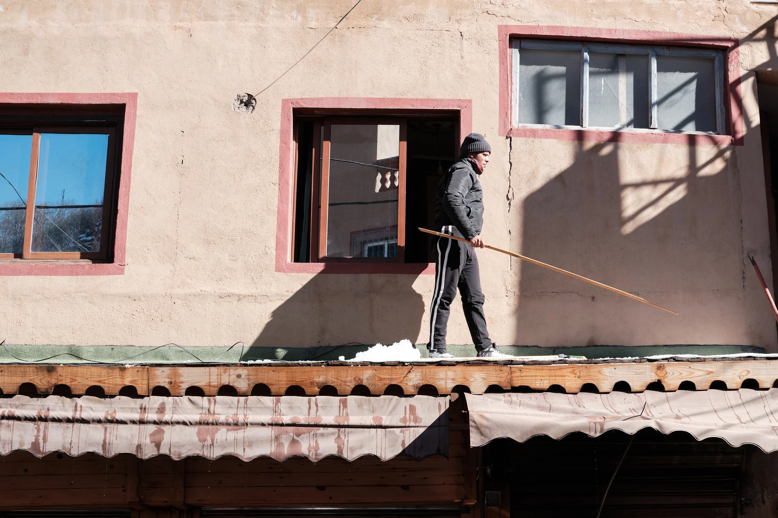 Un uomo che scivola su una superficie ghiacciata davanti a una parete di un edificio con finestre. L'uomo indossa un cappello grigio e una giacca nera.
