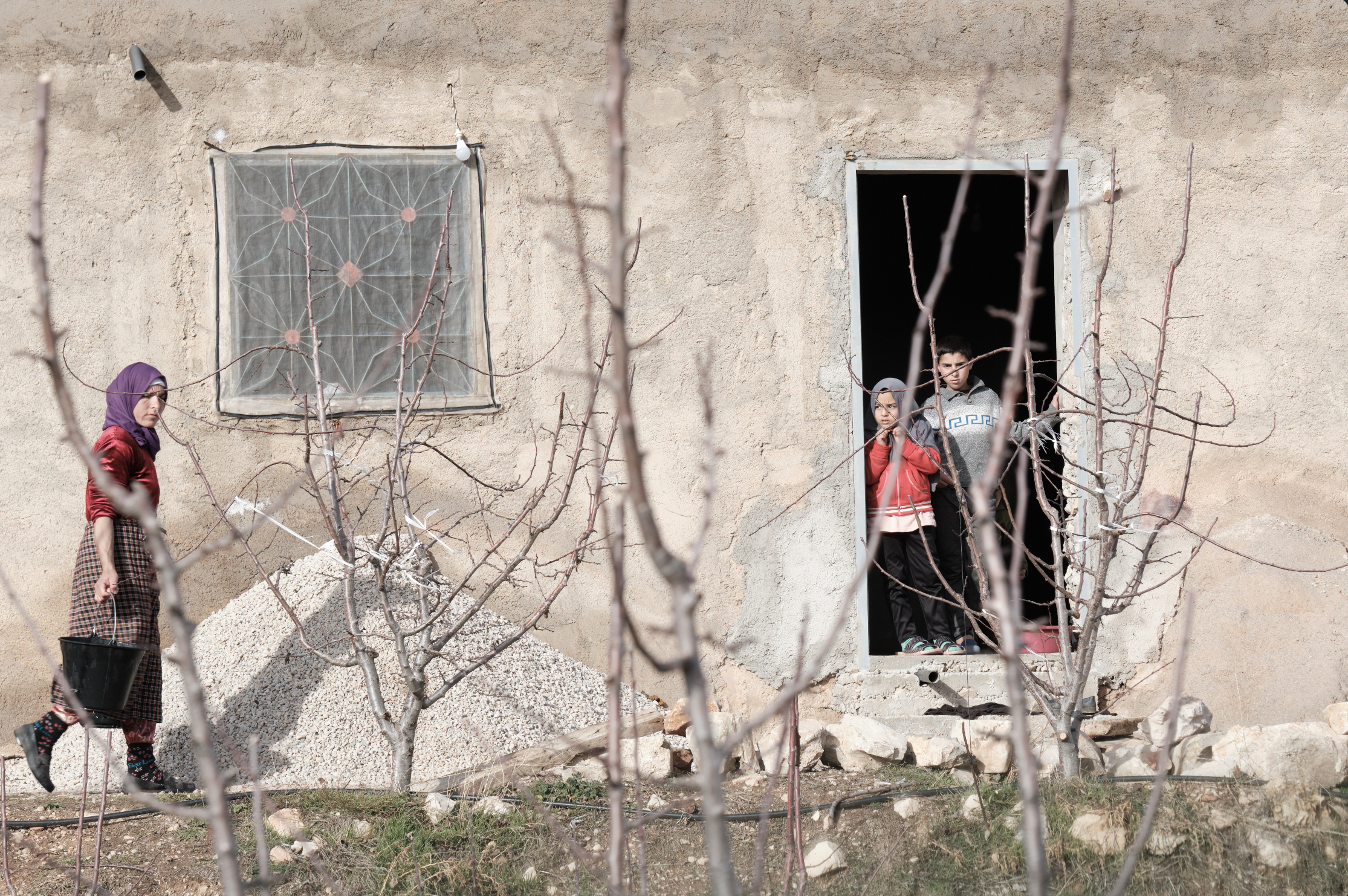 Tre bambini in abiti autunnali vicino a un muro di casa, con alcuni alberi spogli in primo piano.