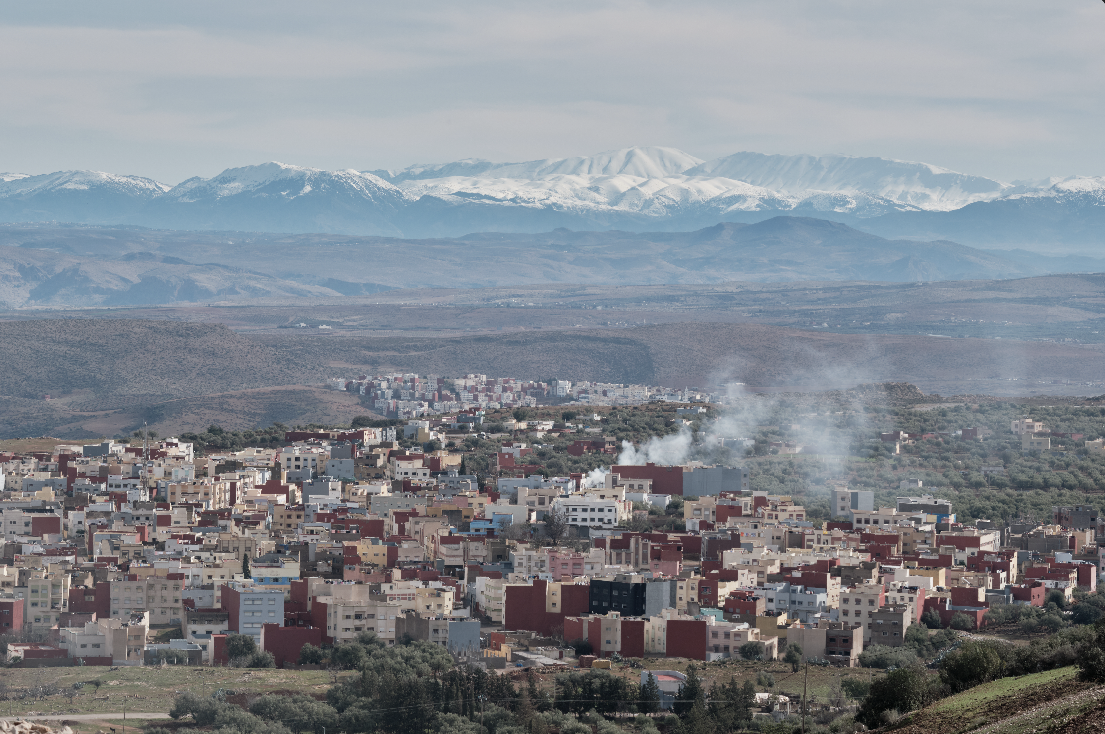 Paesaggio con città di case e palazzi in primo piano, colline sullo sfondo e montagne innevate sotto un cielo nuvoloso.