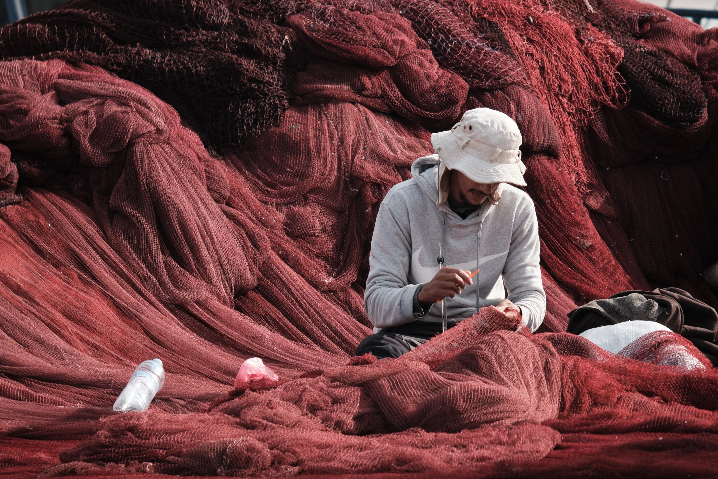 Uomo seduto tra grandi reti di pesca rosse, che lavora con attrezzi, con una bottiglia di plastica sul lato.