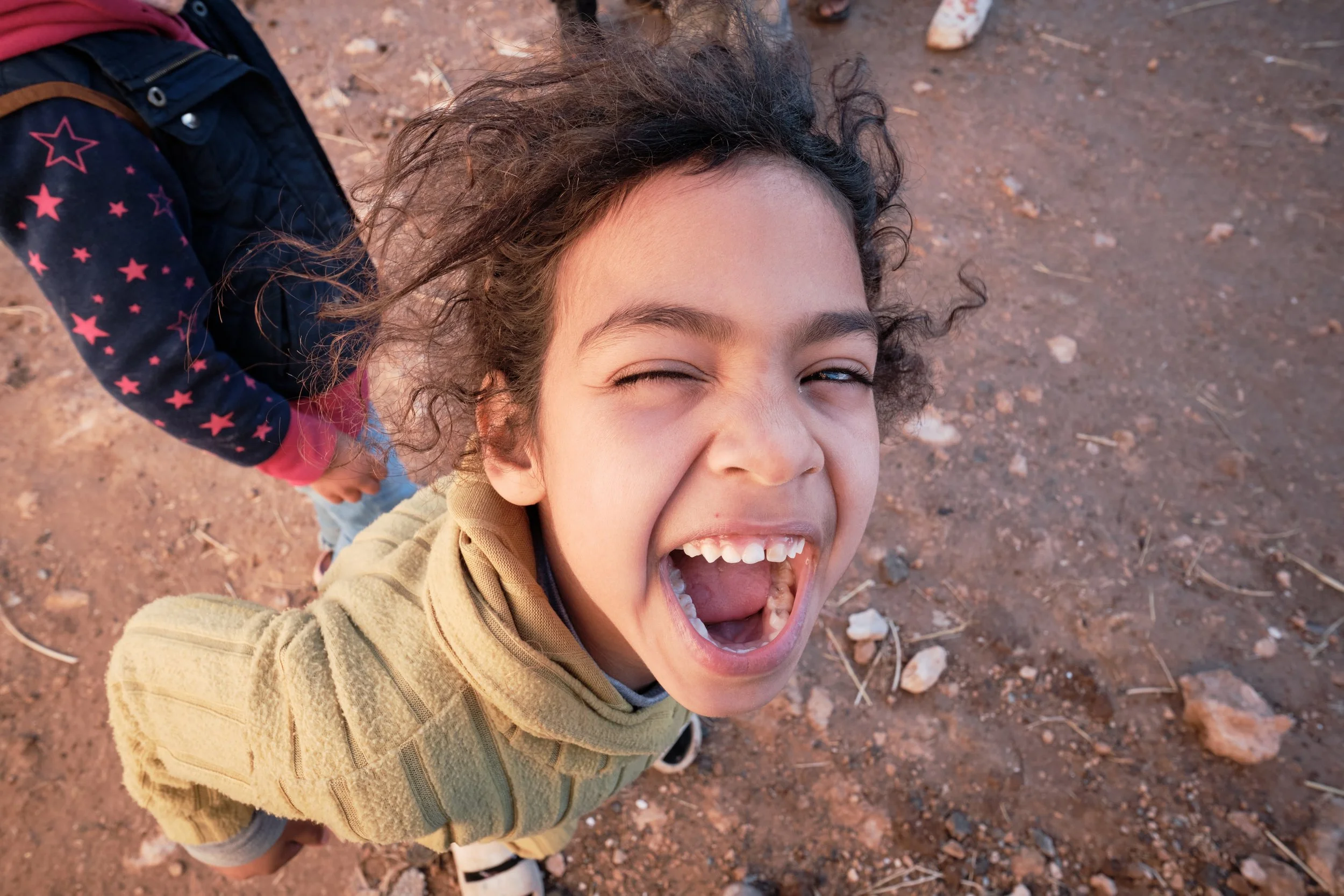 Bambino con capelli ricci, sorriso aperto, che guarda verso la fotocamera, in un ambiente esterno con terra e pietre, che fa un'espressione felice e giocosa.