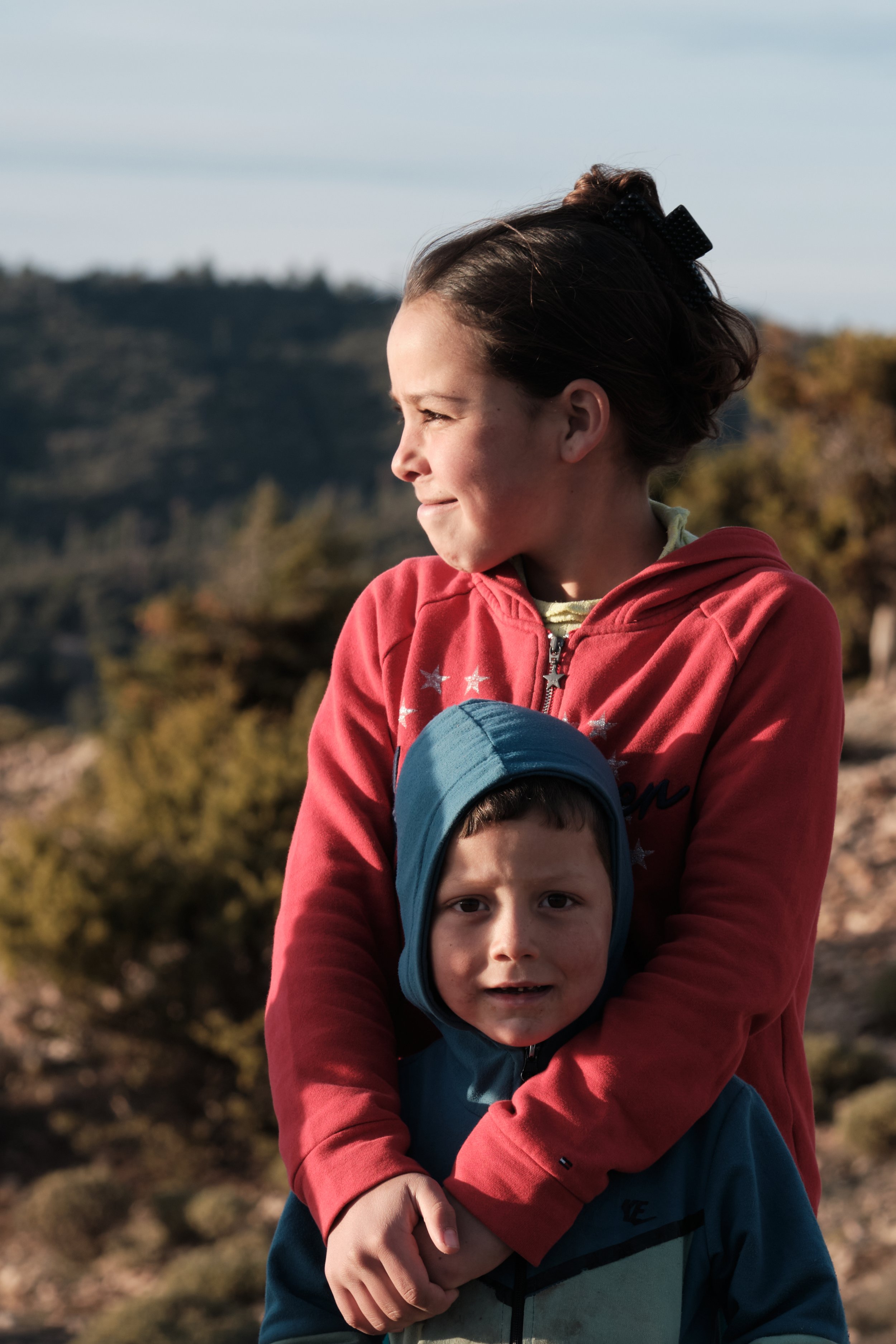 Una ragazza con i capelli raccolti, indossa una felpa rossa, abbraccia un bambino con cappello blu e felpa blu, in un ambiente all'aperto con natura e colline sullo sfondo.