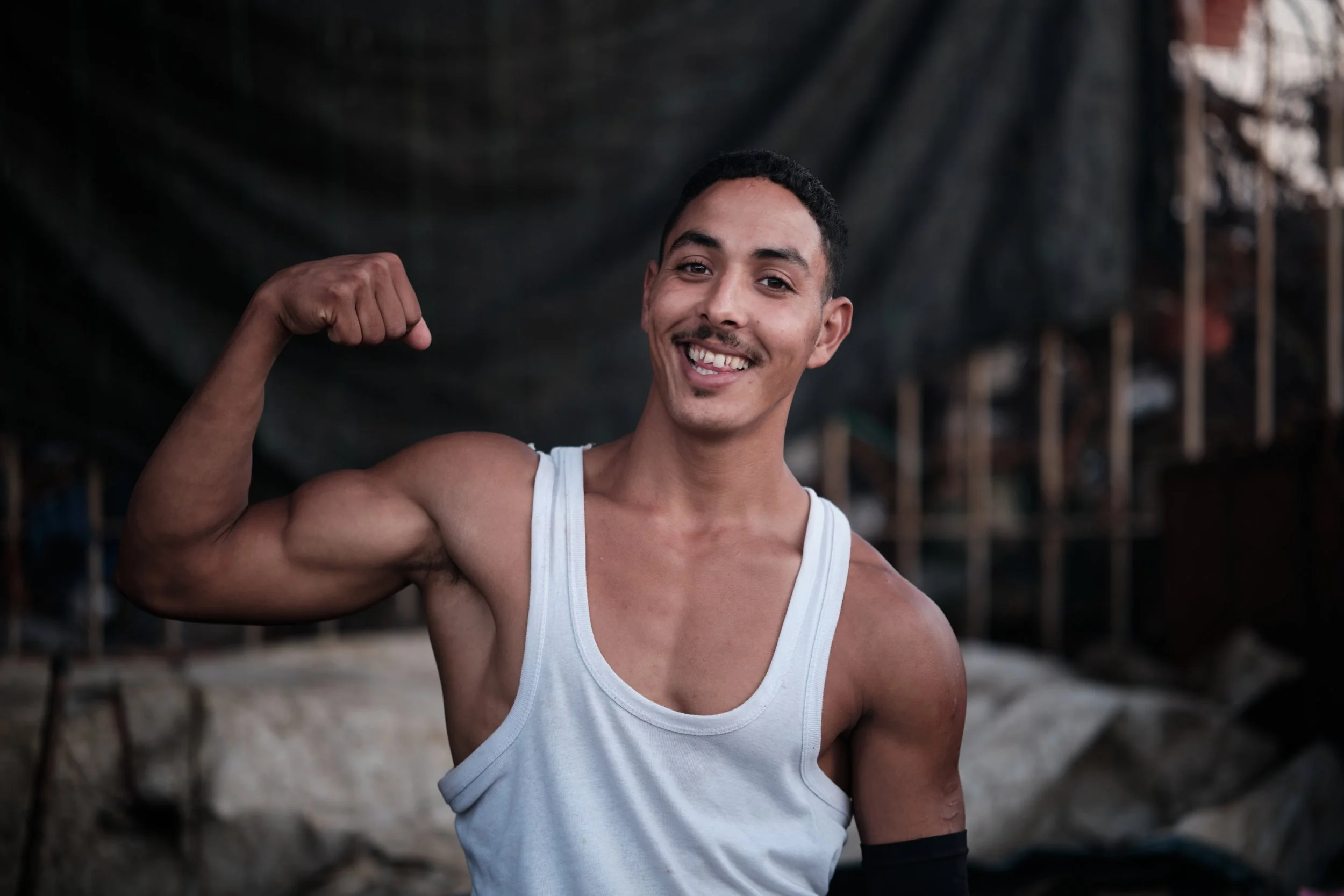 Giovane uomo con capelli corti, sorriso e muscoli visibili, che si frega il bicipite mentre indossa una canotta bianca, ambientato in un luogo aperto.