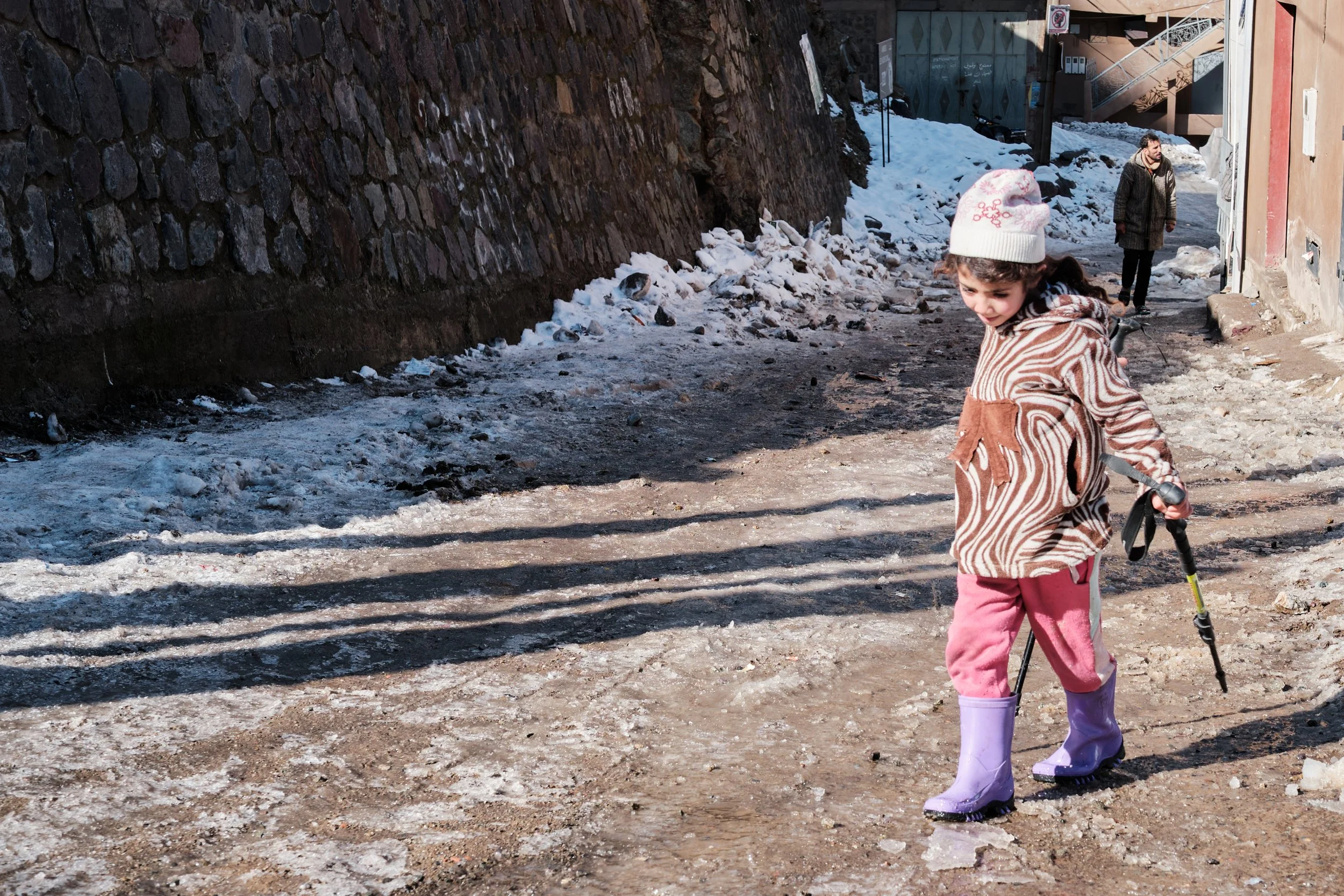 Due bambini che giocano nella neve in una strada di montagna, uno con giacca a strisce rosa e marrone, e l'altro più lontano con cappotto a quadri. Sullo sfondo, un muro in pietra e una casa con scale e uscite sulle pareti