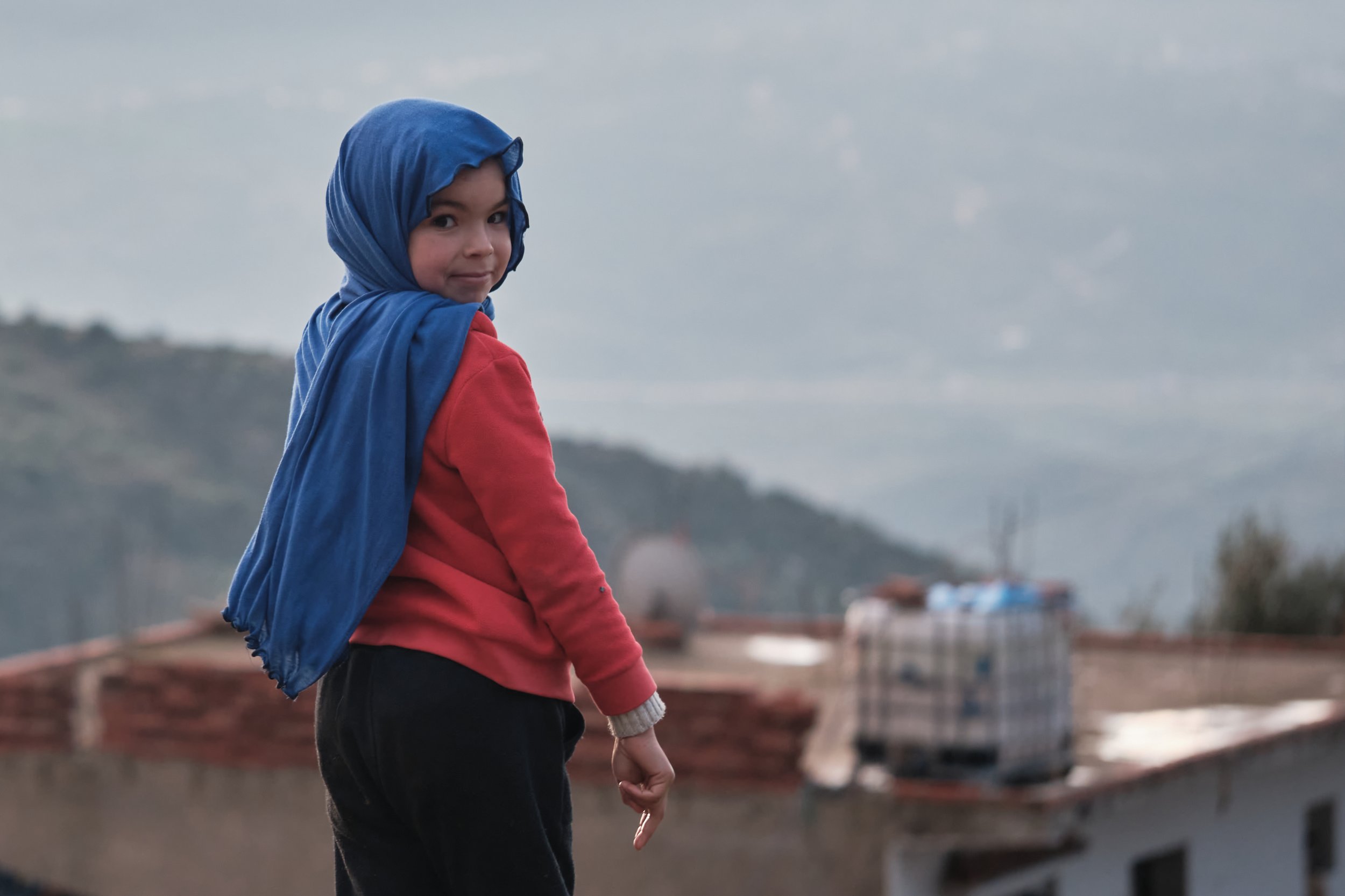 Bambina con cappuccio blu indossa una maglietta rossa e pantaloni neri, guarda verso l'obiettivo con uno sfondo di cielo grigio e paesaggio urbano.