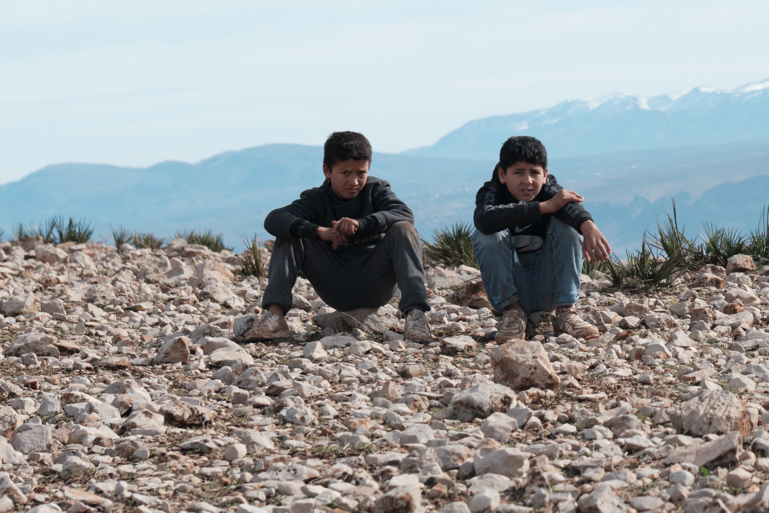Due bambini seduti su un terreno di sassi in un'area aperta con montagne sullo sfondo, uno dei bambini sembra triste o pensieroso.
