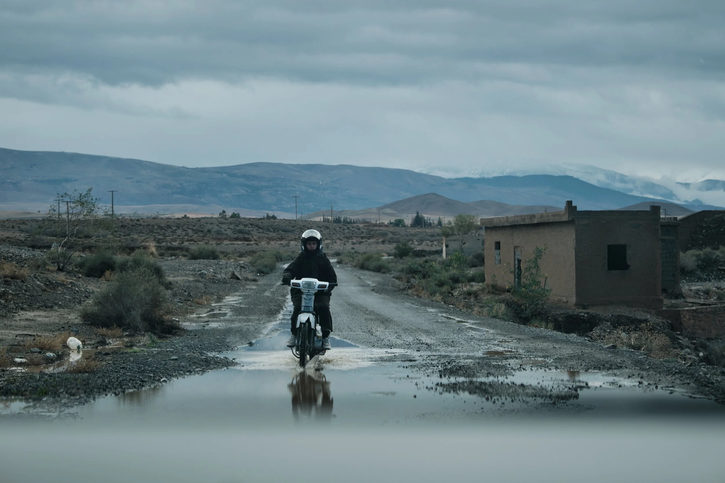 Un motociclista attraversa una strada sterrata in un paesaggio desertico con montagne sullo sfondo. Il cielo è nuvoloso e ci sono alcune case abbandonate ai lati della strada.