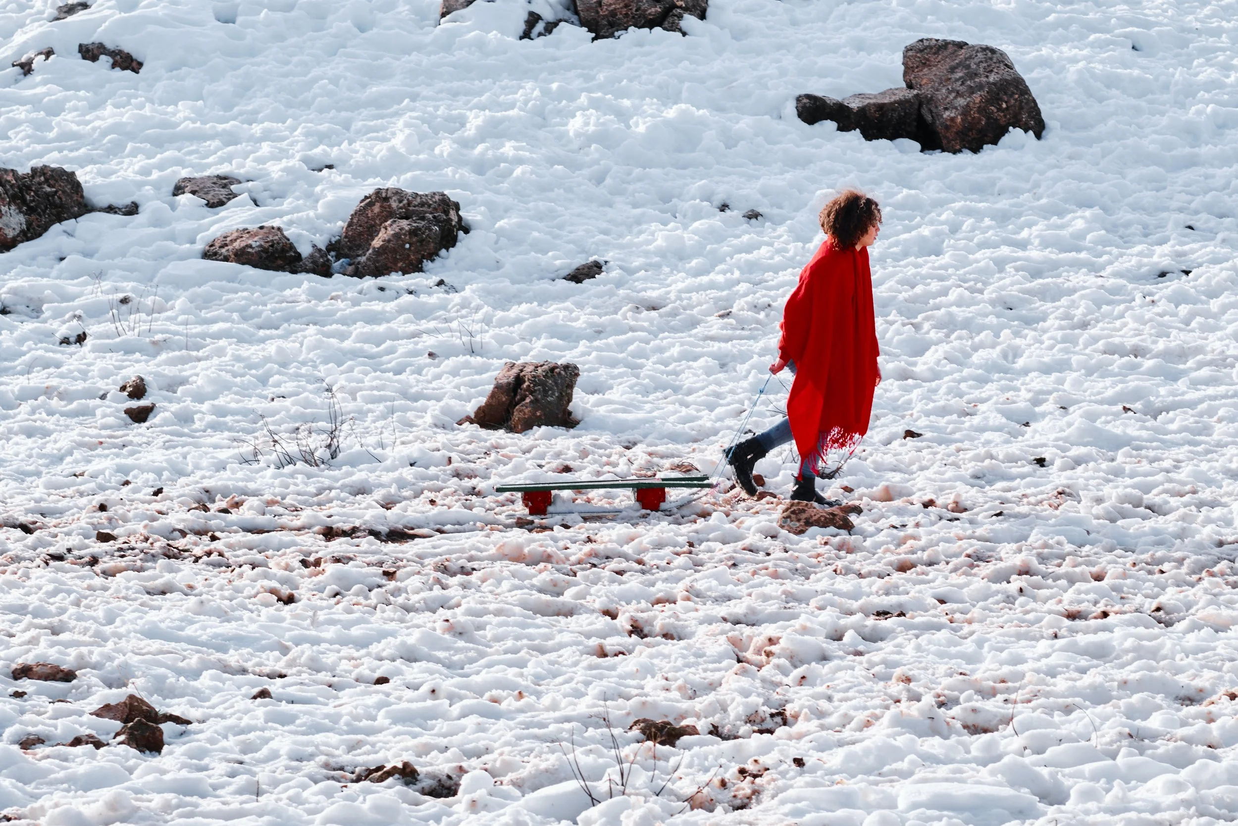 Una donna con capelli ricci e vestito rosso cammina sulla neve con un bastone, trascinando una slitta dietro di sé.