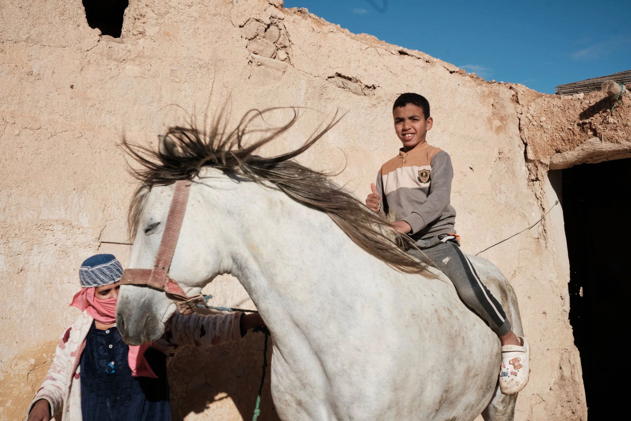 Un bambino in sella a un cavallo bianco con una persona più piccola vicino che lo tiene. Sullo sfondo, un muro di pietra e un cielo azzurro con alcune nuvole.