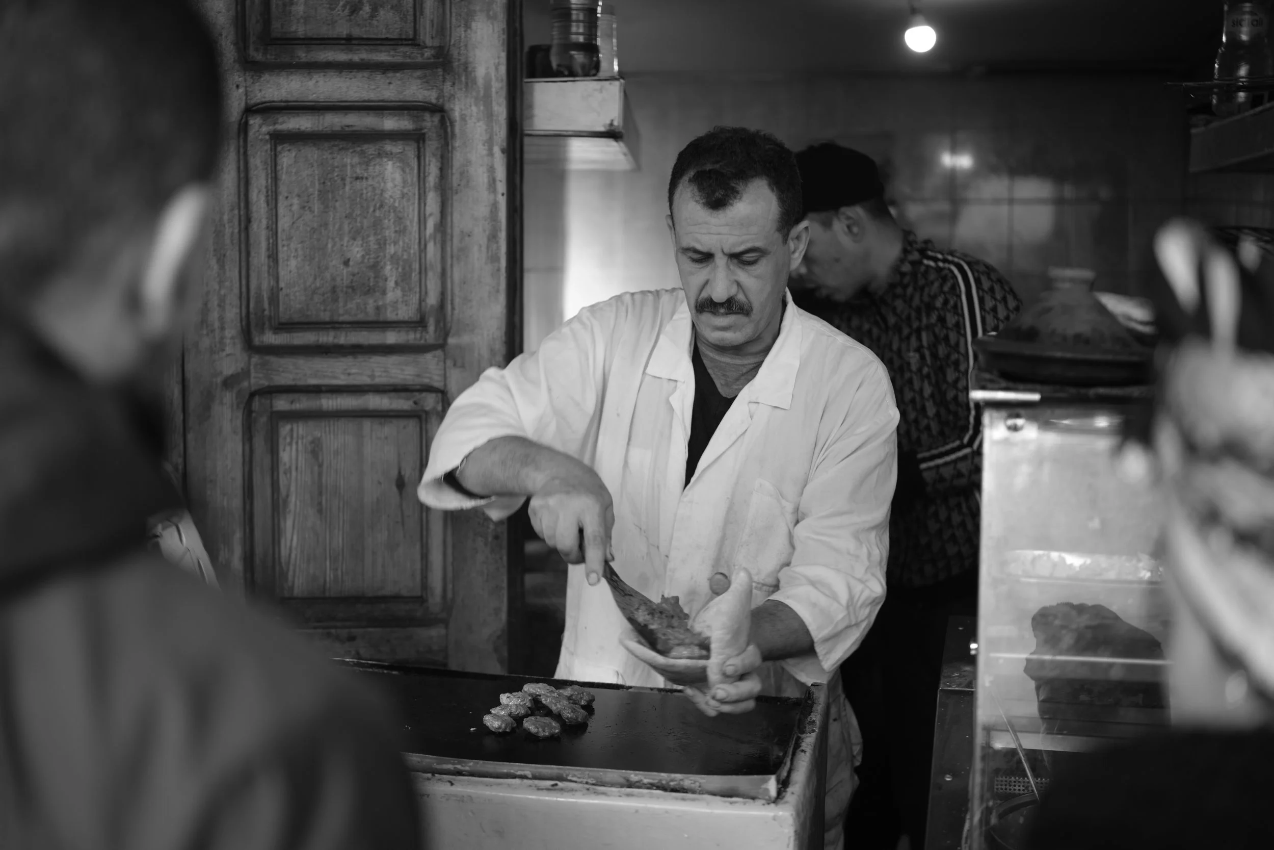 Uno chef indossa una giacca bianca mentre prepara carne su un piatto di cucina in un ristorante, con altri persone sullo sfondo.