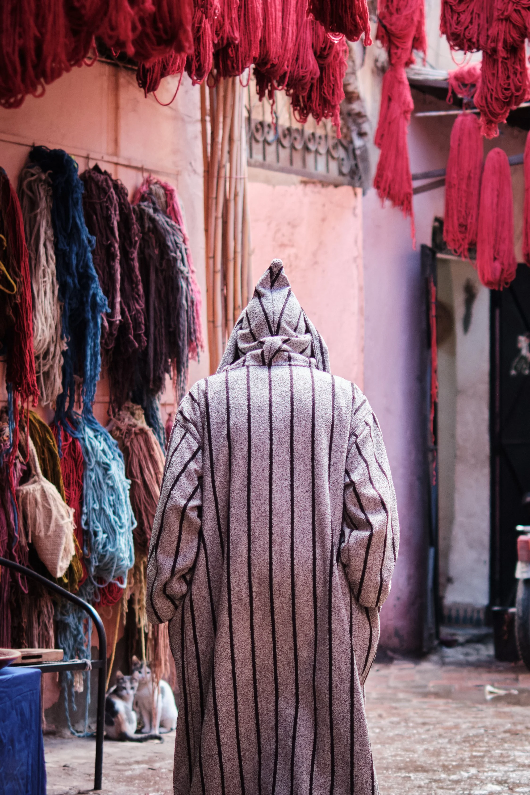 Donna con cappotto a righe che cammina in una piazza, con tessuti colorati appesi lungo le pareti e cani in piedi in fondo.