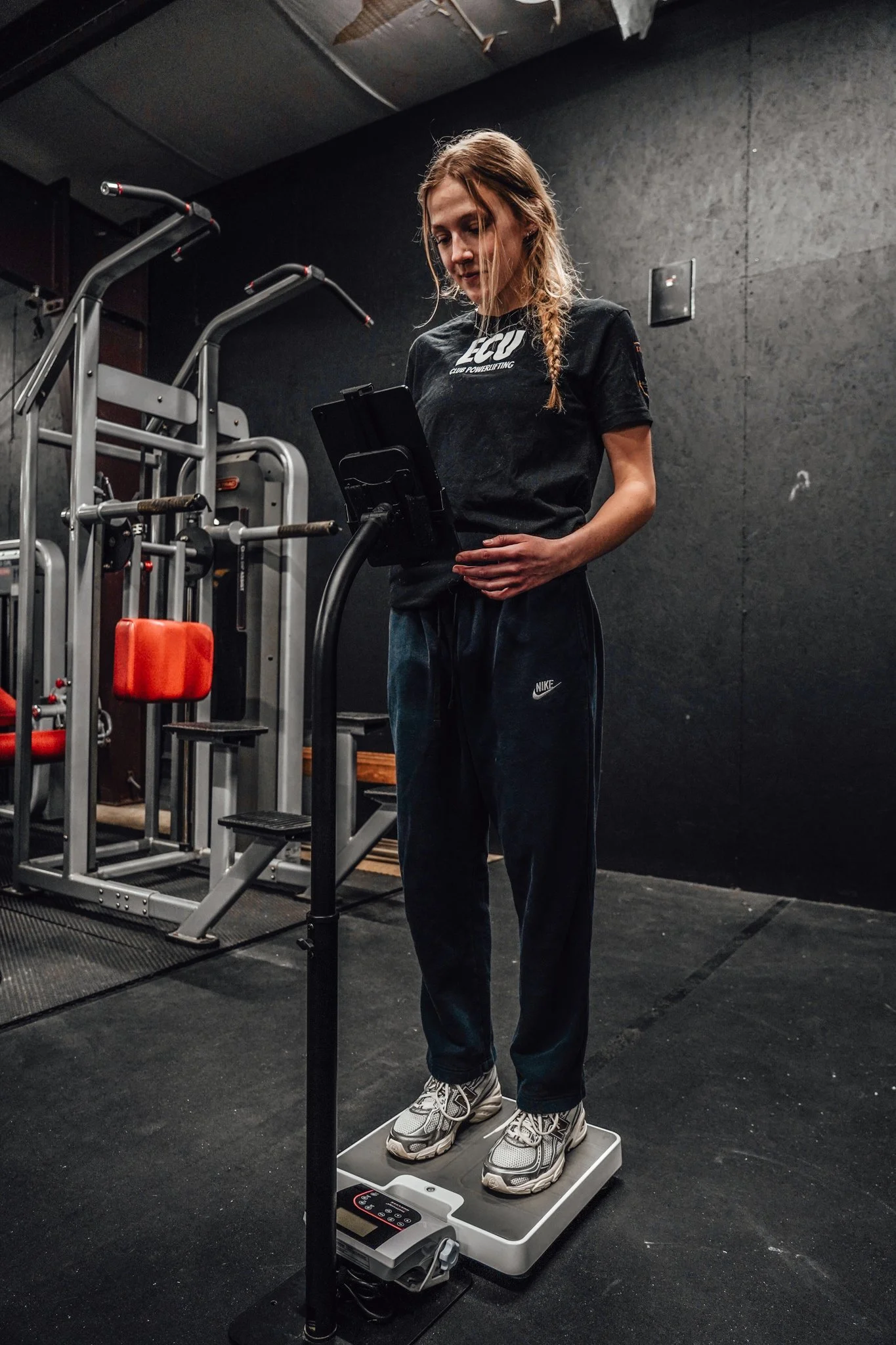A woman with long hair wearing a black T-shirt and black Nike pants stands on a weighing scale in a gym or fitness studio. She appears to be measuring her weight digitally, with some gym equipment visible in the background.