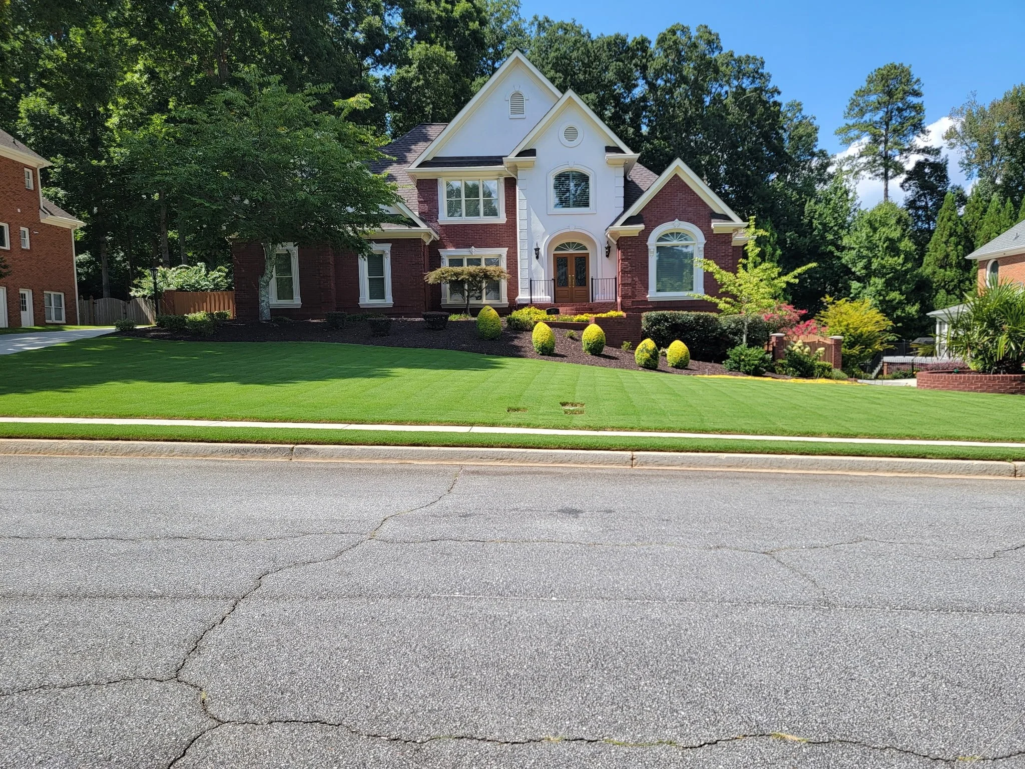 A front view of a large, two-story house with a combination of red brick and white exterior trim, surrounded by a well-maintained lawn and lush green trees, located on a suburban street.