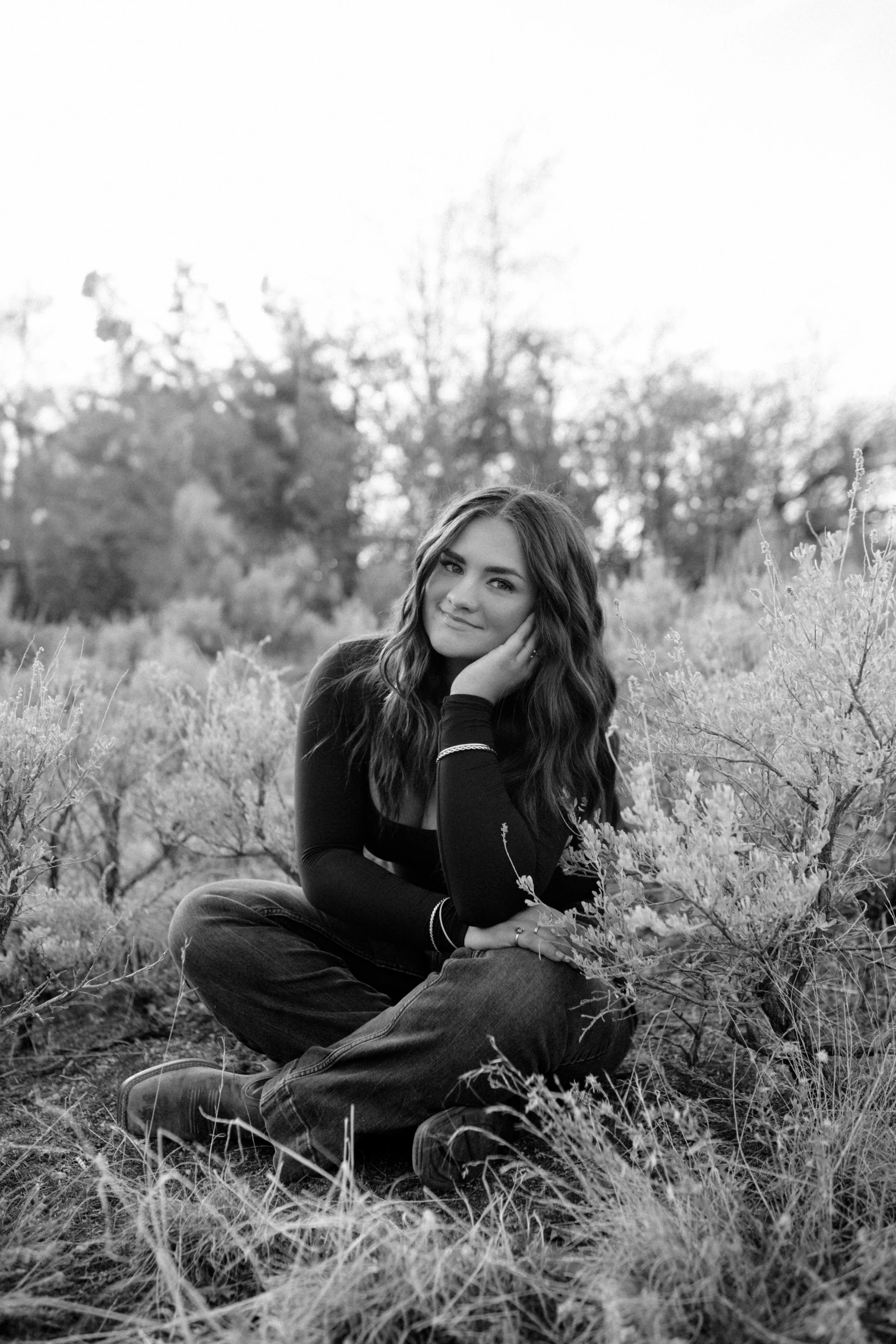 A young woman with long hair, wearing a dark long-sleeve shirt and jeans, sitting cross-legged on grass in a field of small plants, smiling and resting her face on her hand, with trees and bare branches in the background, in black and white.