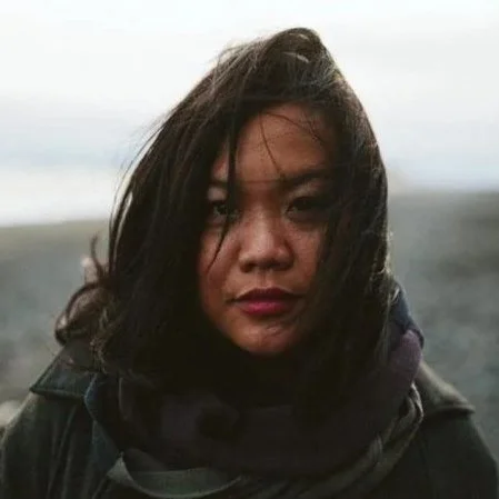 Woman with dark hair and windblown hair standing outdoors near a beach or shoreline.