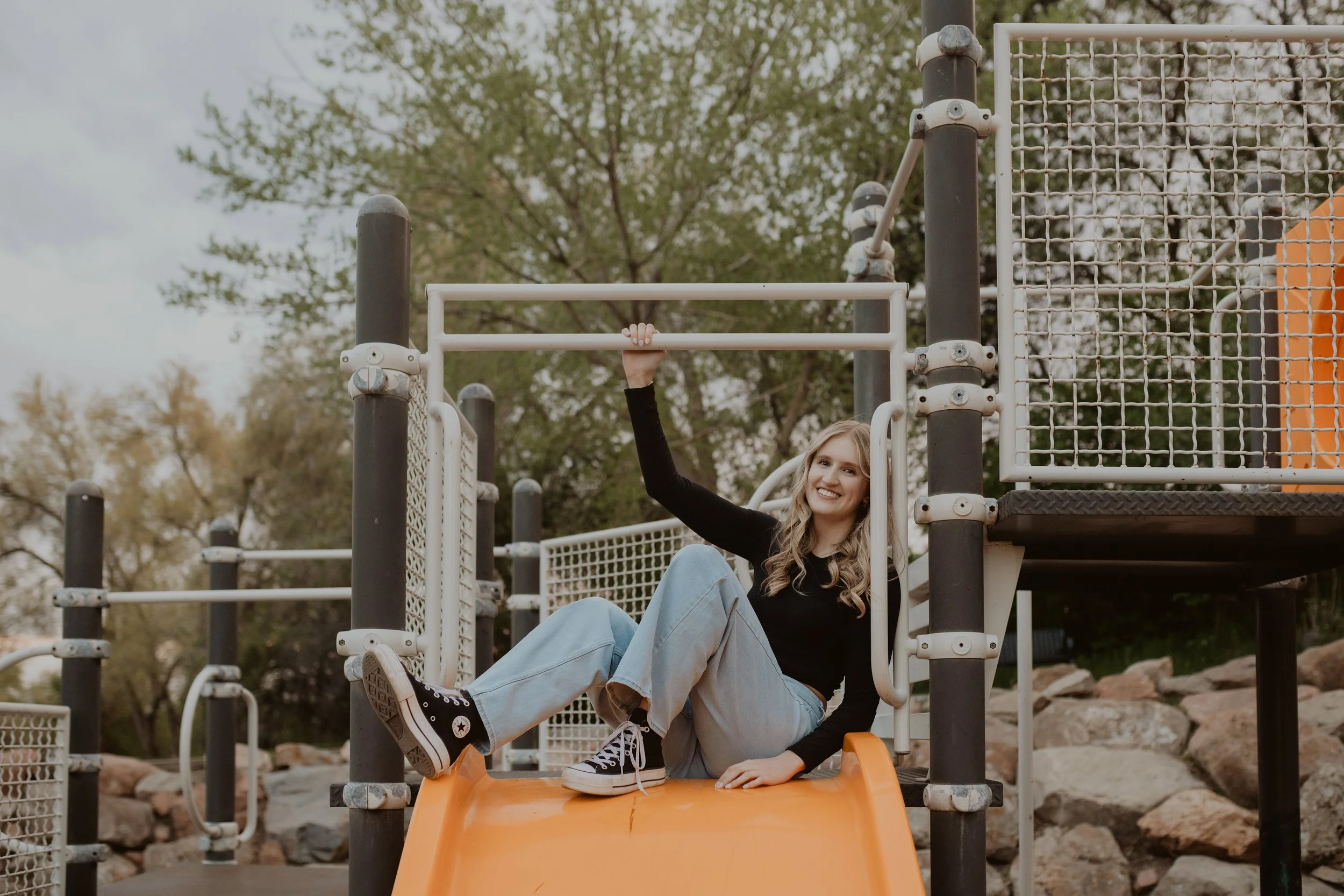 Young woman sitting on a slide at a playground, smiling, with one arm raised, surrounded by playground equipment and trees in the background.