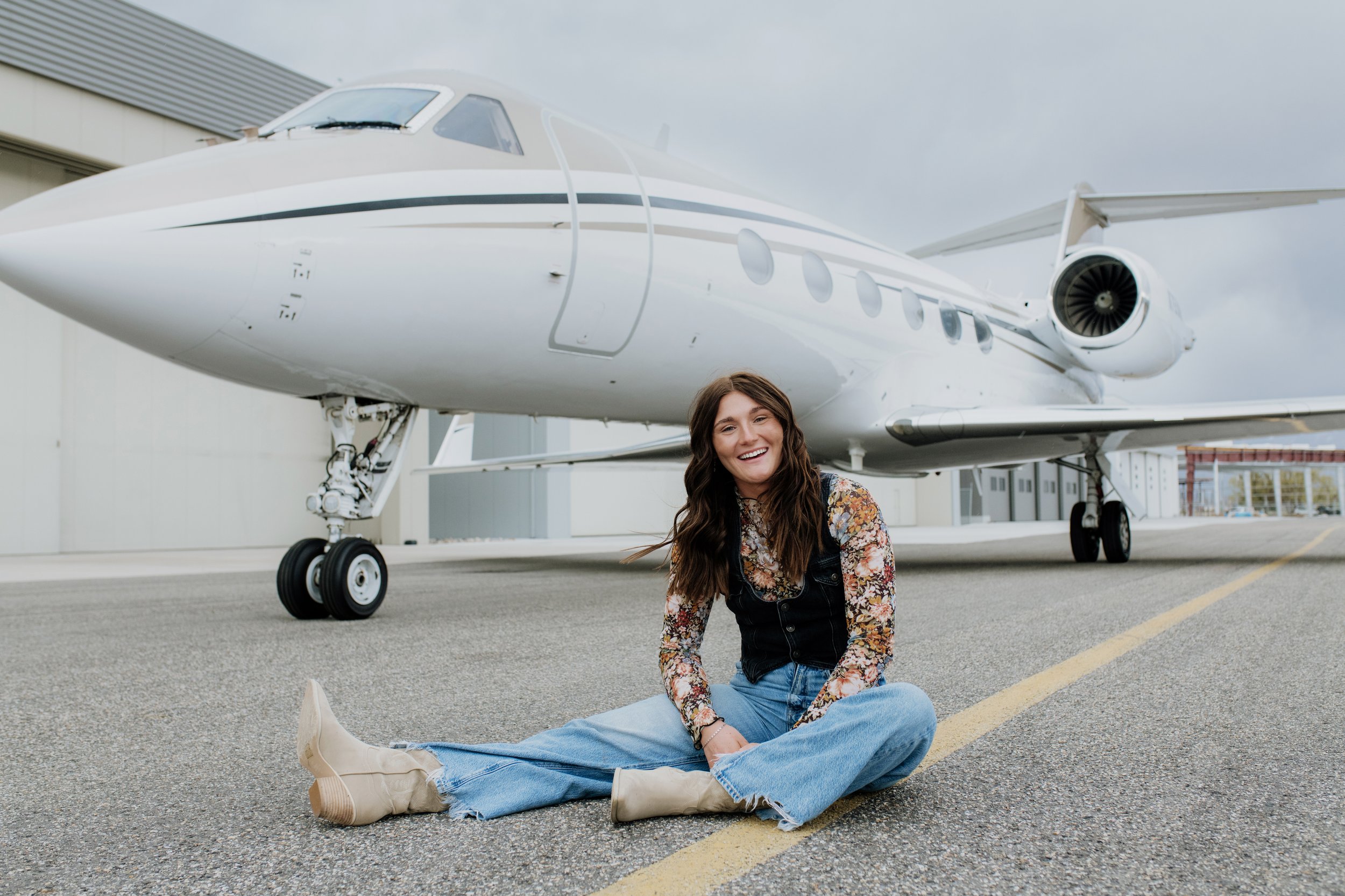 A woman sitting on the ground in front of a private jet airplane, smiling and looking happy.