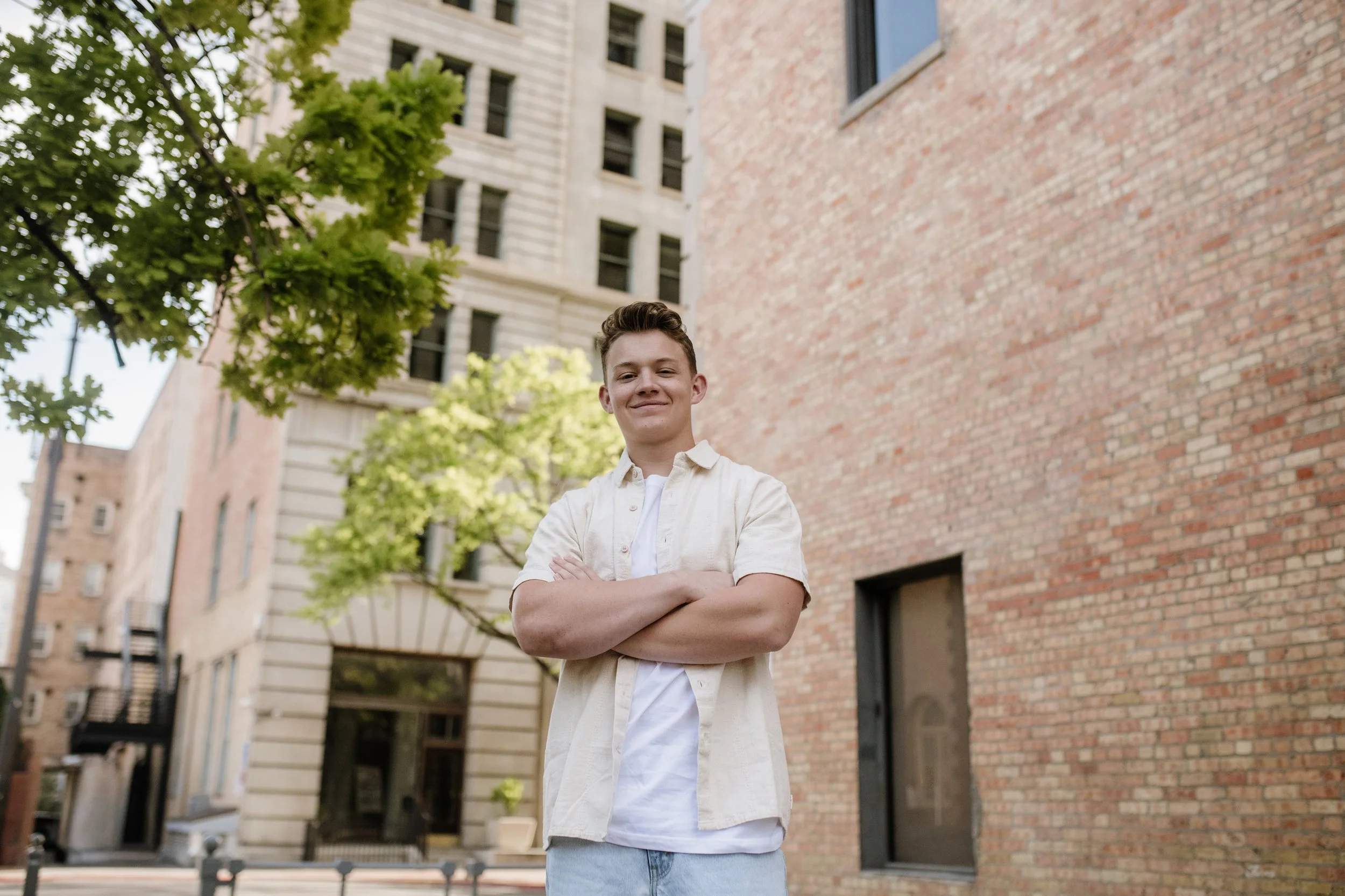 A young man with short hair standing outdoors with arms crossed, smiling in front of a brick building and trees.