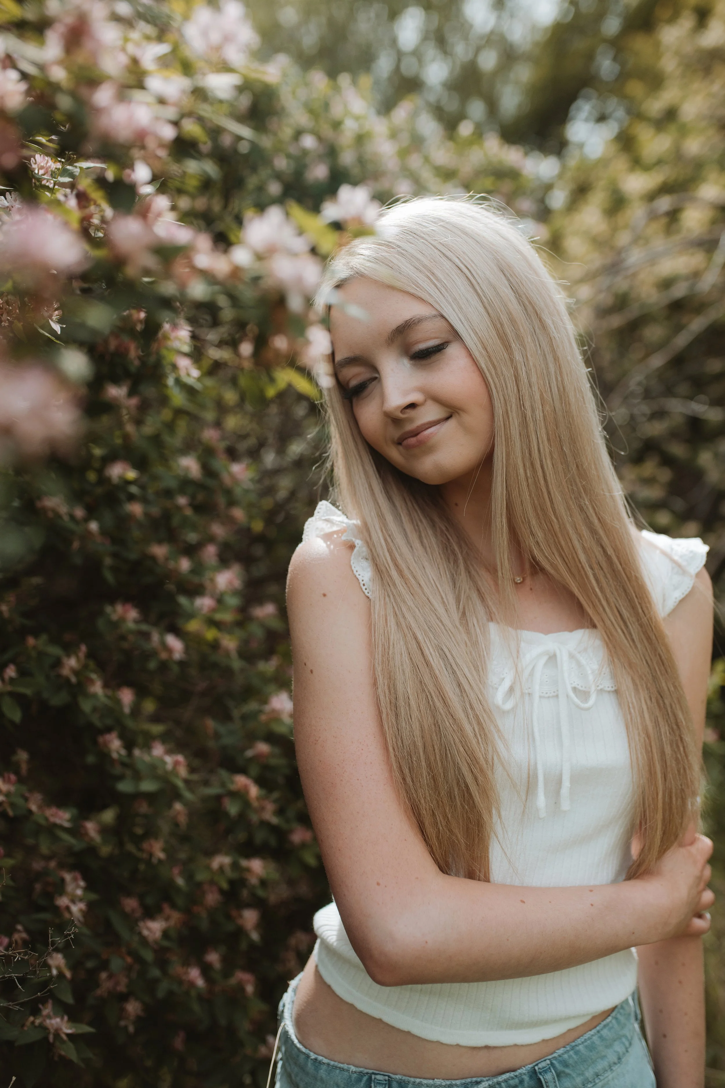 A young woman with long blonde hair, closed eyes, and a smile, standing outdoors near flowering bushes.