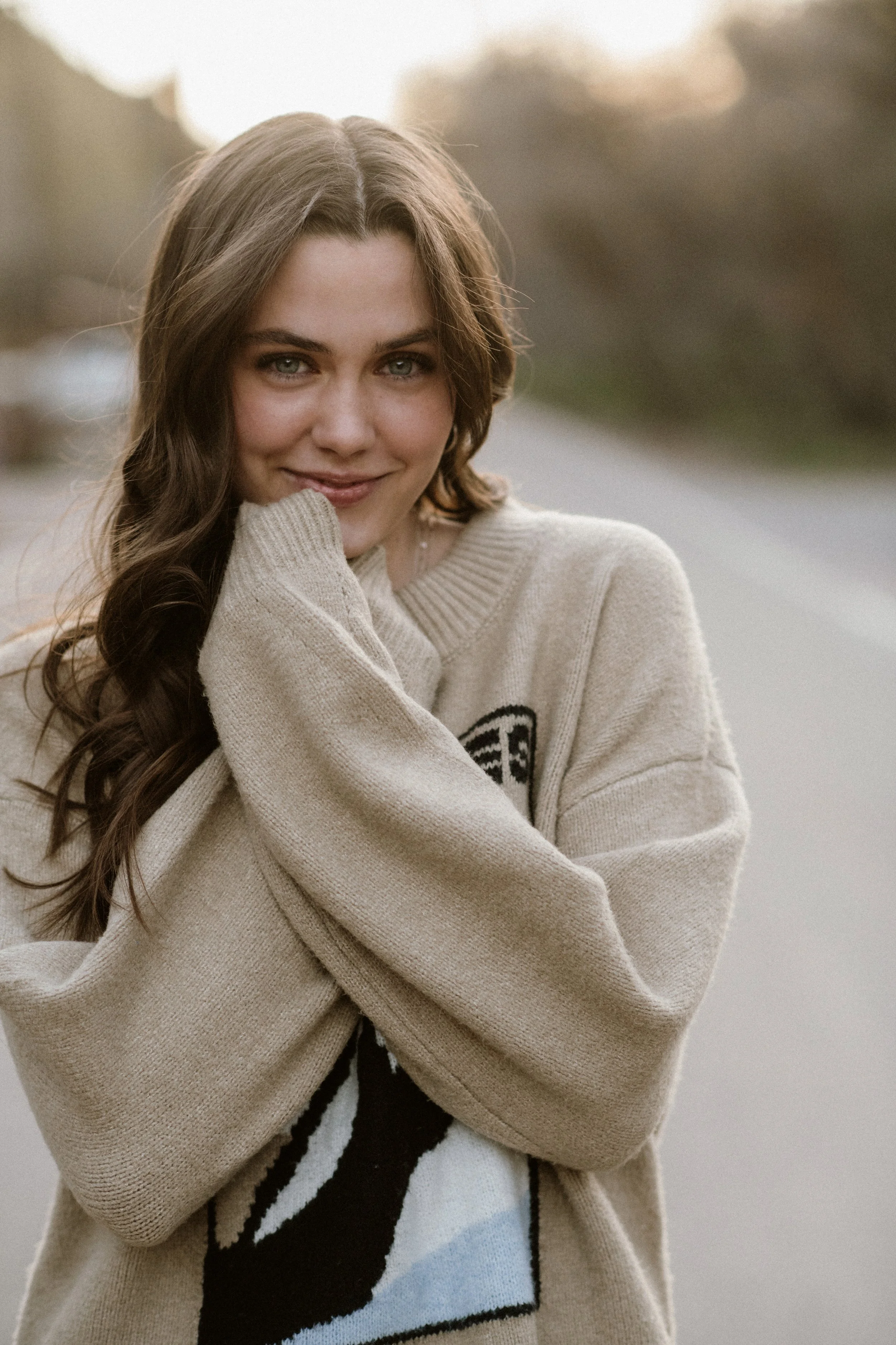 Young woman with long brown hair and blue eyes, wearing a beige sweater and a white top with a black graphic, smiling and looking at the camera outdoors.
