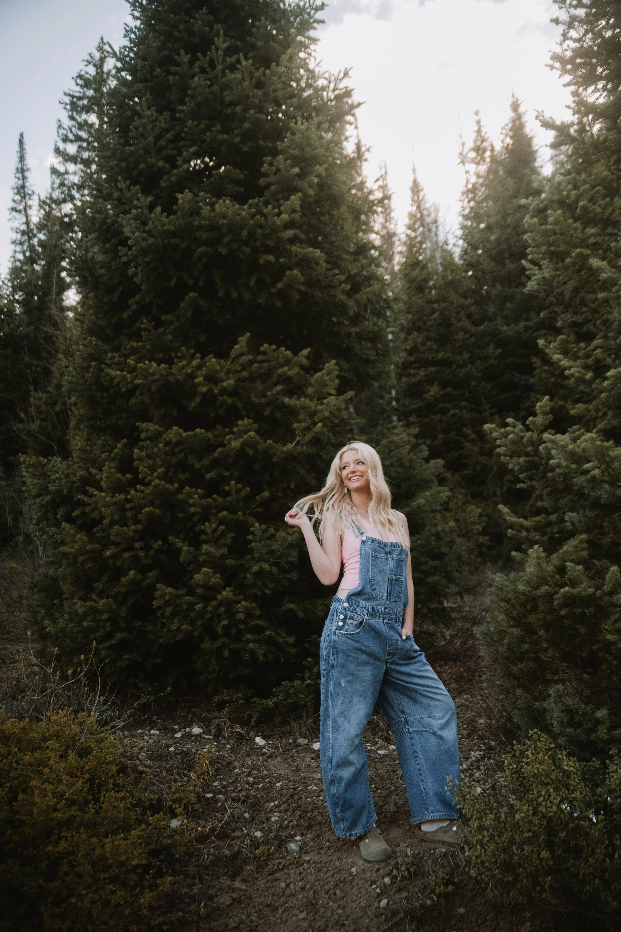 Young woman with long blonde hair standing outdoors in front of pine trees, wearing denim overalls, a pink tank top, and beige shoes, smiling and looking to her right.