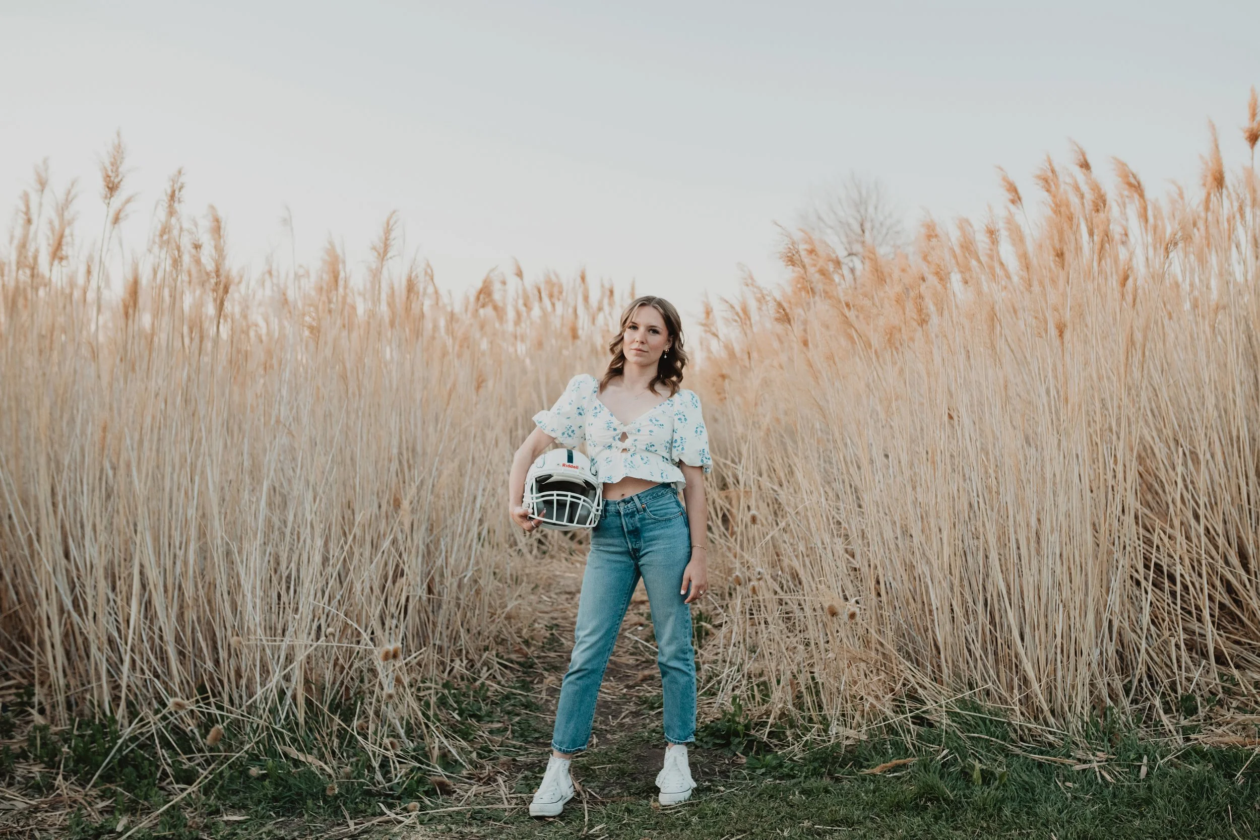 Young woman standing in a field of tall, beige grass, holding a white helmet, wearing high-waisted jeans, a floral crop top, and white sneakers.