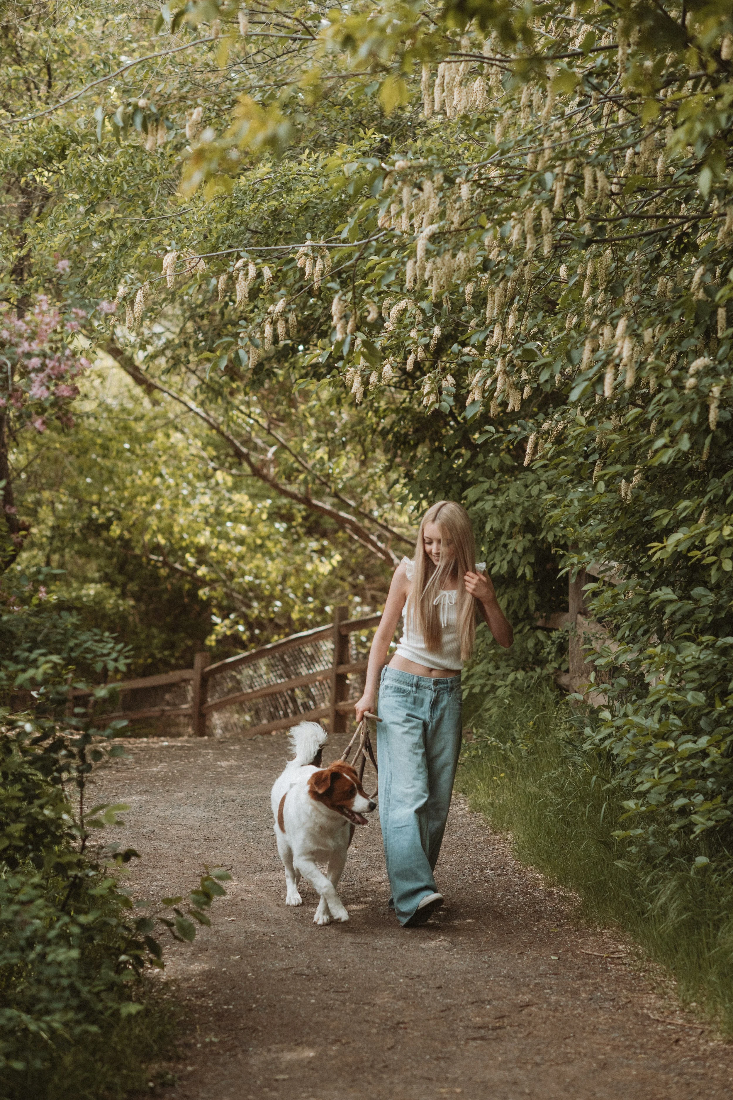 A young woman with long blond hair wearing a white sleeveless top and baggy jeans walking her dog on a dirt path surrounded by lush green foliage and trees, with a wooden fence on the side.