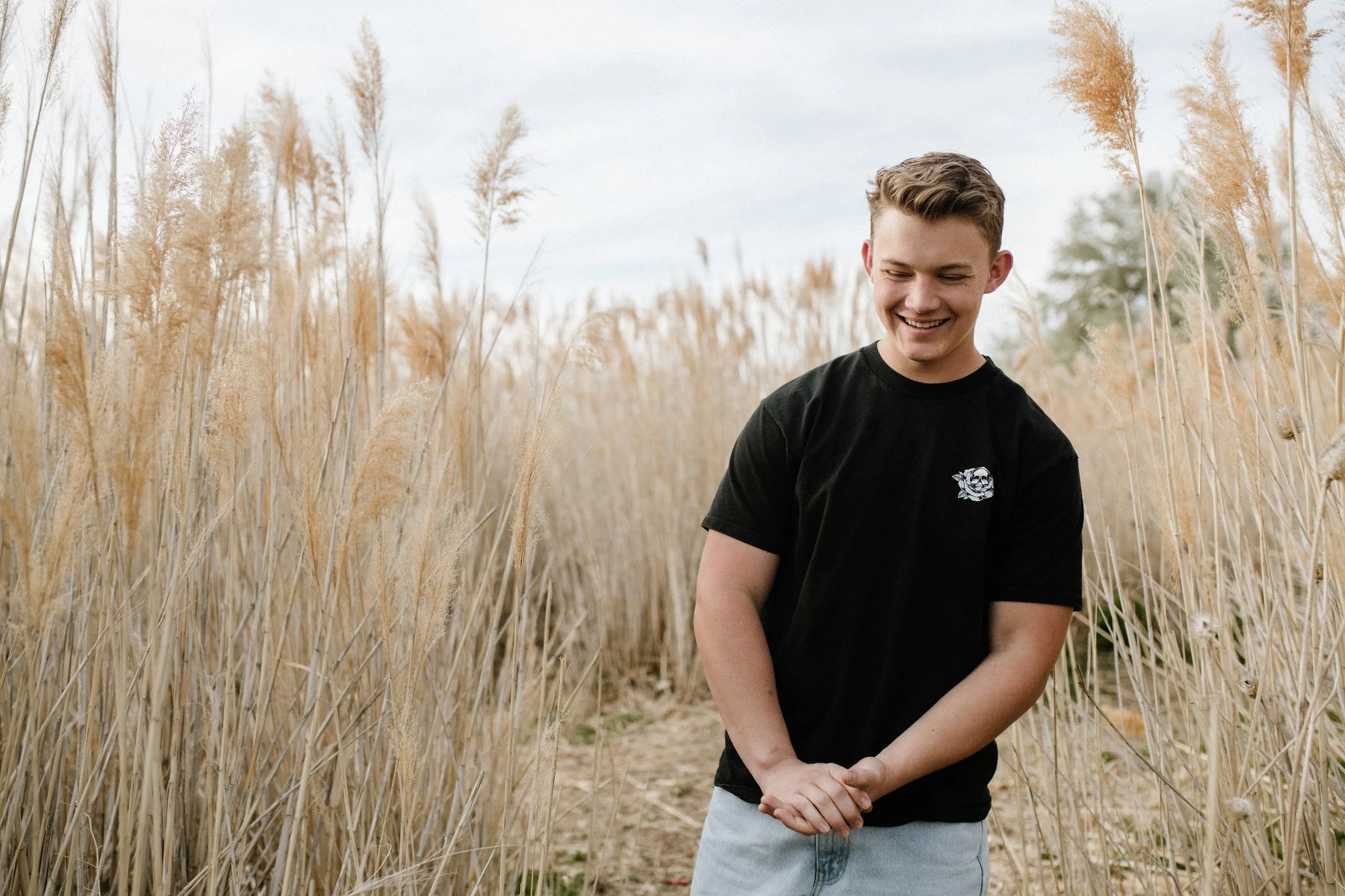 A young man standing in a field of tall, golden grasses, smiling and looking down, wearing a black t-shirt and light-colored jeans.