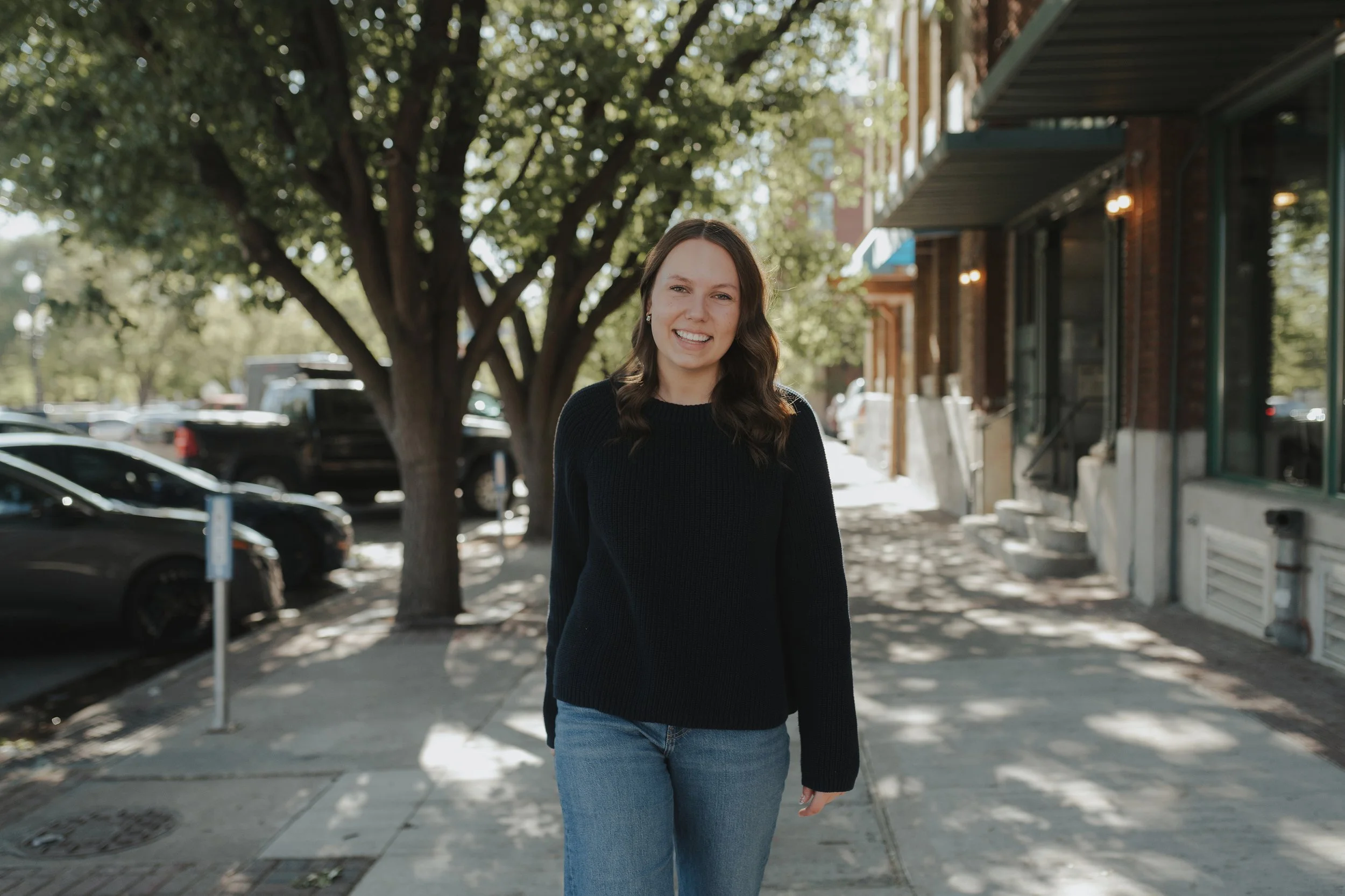 A young woman with brown hair and wearing a black sweater walks along a city sidewalk during the daytime. There are trees and parked cars on her left and storefronts on her right.