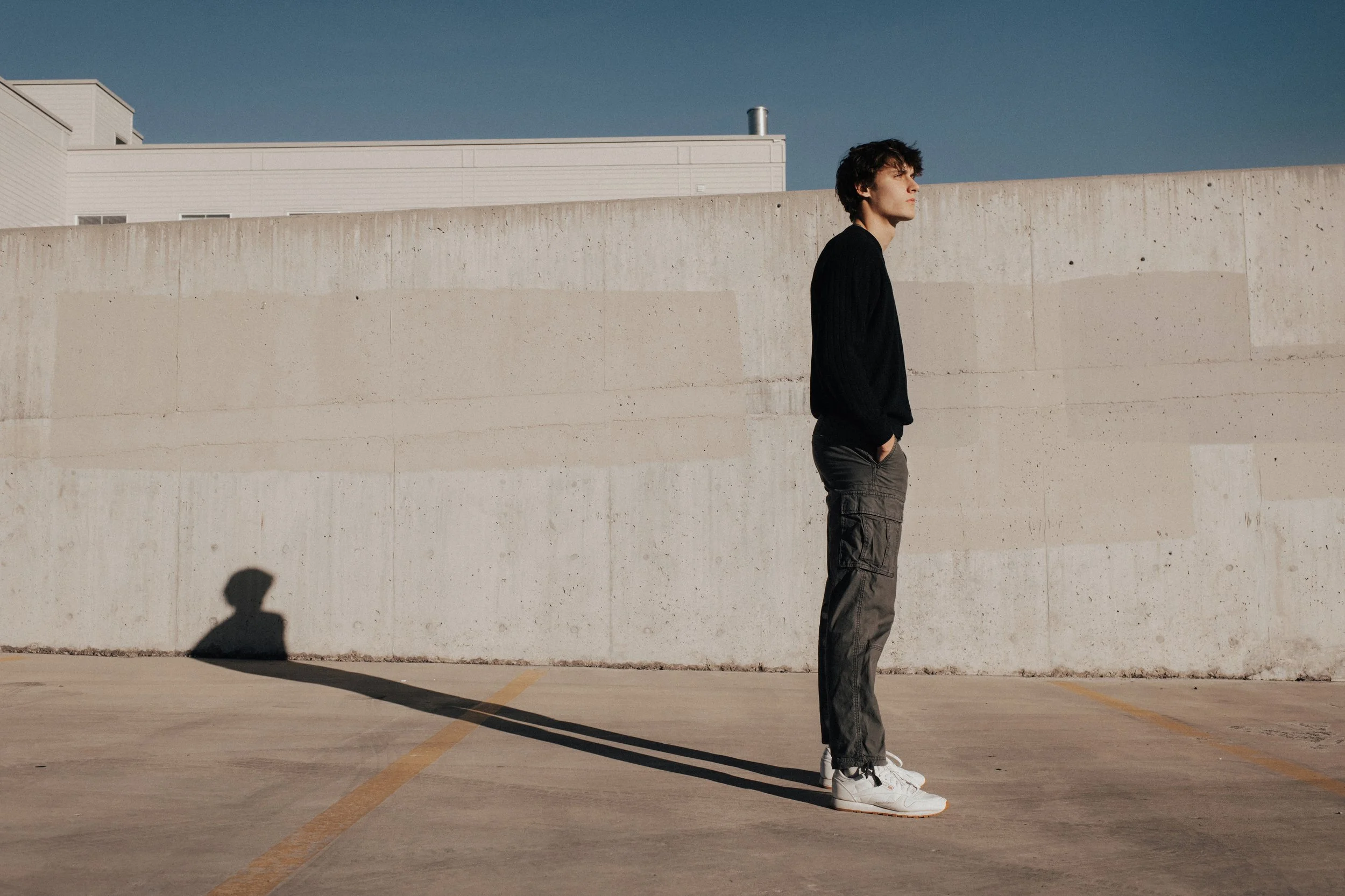 Young man with dark hair and light skin wearing a black sweater, cargo pants, and white sneakers standing in an empty parking lot near a large concrete wall, with his shadow cast on the ground and a modern building in the background under a clear blu