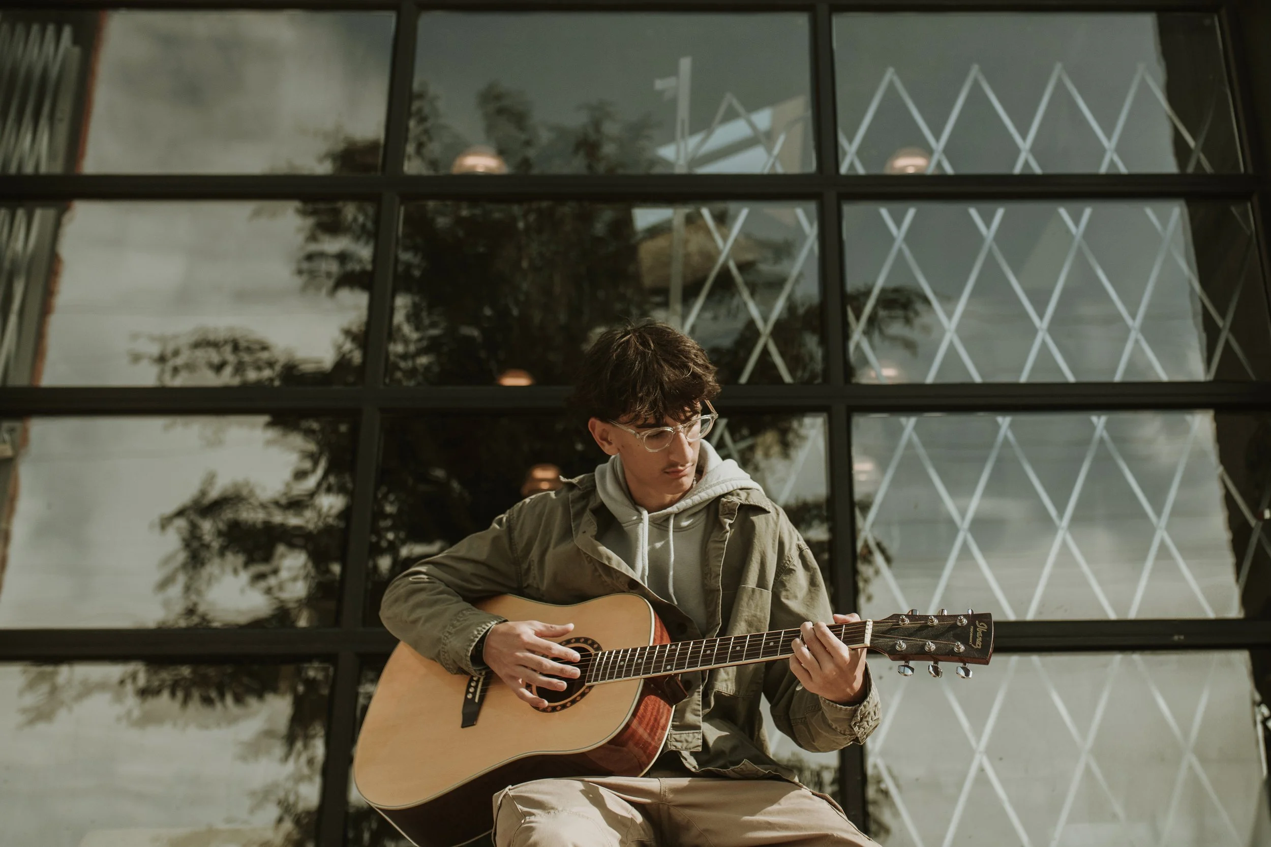 Senior boy with glasses and dark, wavy hair is sitting outside playing an acoustic guitawearing a beige jacket and a gray hoodie. Behind is a large modern glass building with reflections of trees and an overcast sky.