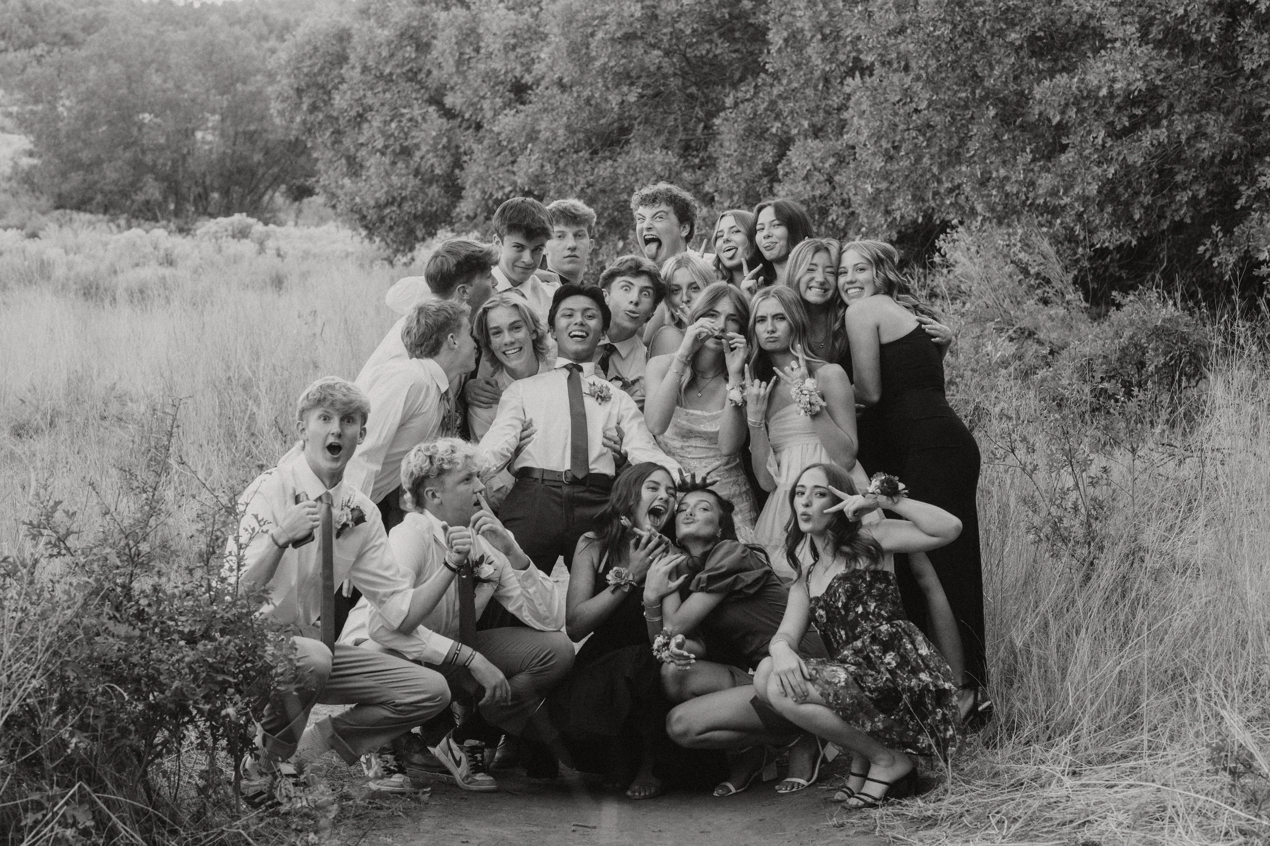 A group of young people gathered outdoors in a field near trees, posing for a photo. They are making playful faces and hand gestures, and some are wearing formal or semi-formal clothing, suggesting a celebration or special event.