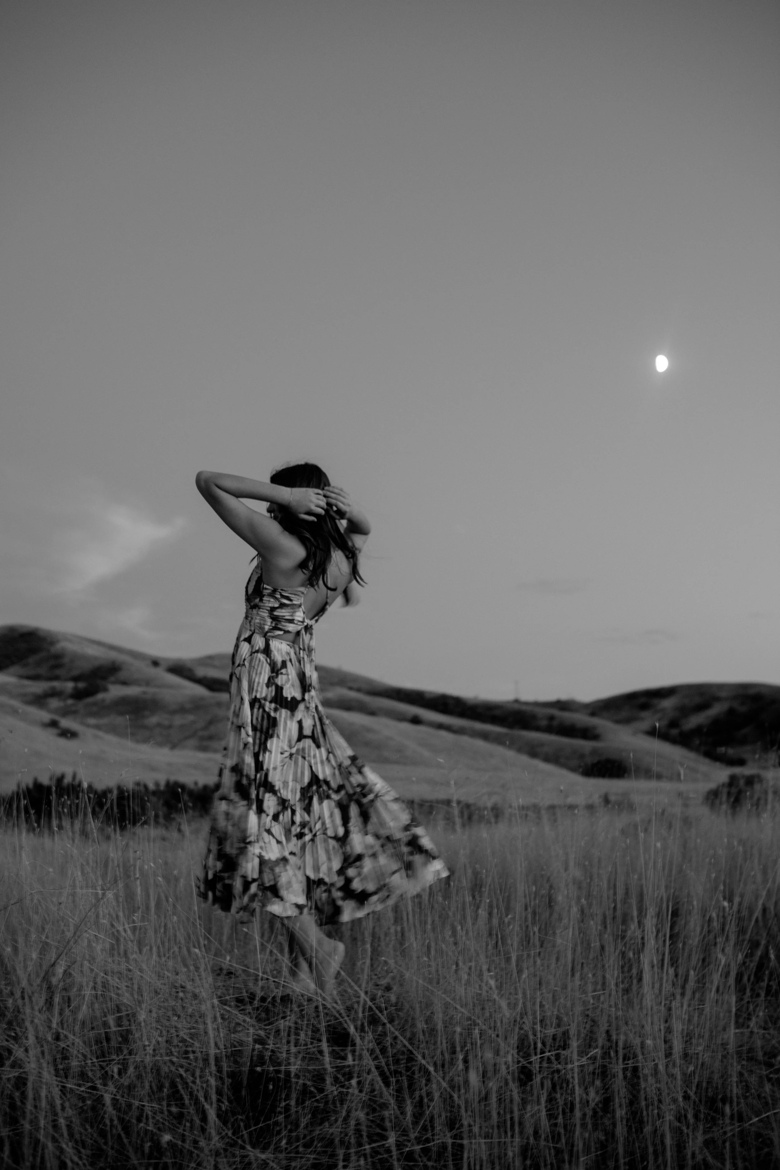 A woman in a patterned dress standing in a field of tall grass, holding her head with both hands, with rolling hills in the background and the moon visible in the evening sky.