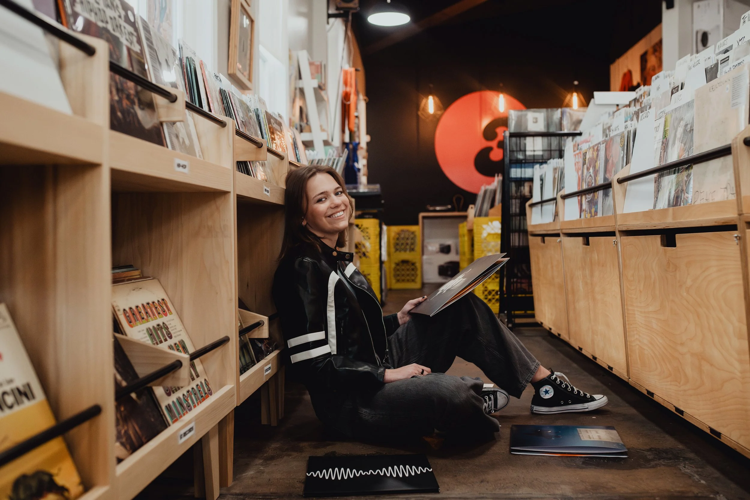 A young woman with brown hair smiling while sitting on the floor of a bookstore, leaning against wooden shelves filled with vinyl records and magazines. She is wearing a black leather jacket, gray pants, and black sneakers. There is a large red logo 