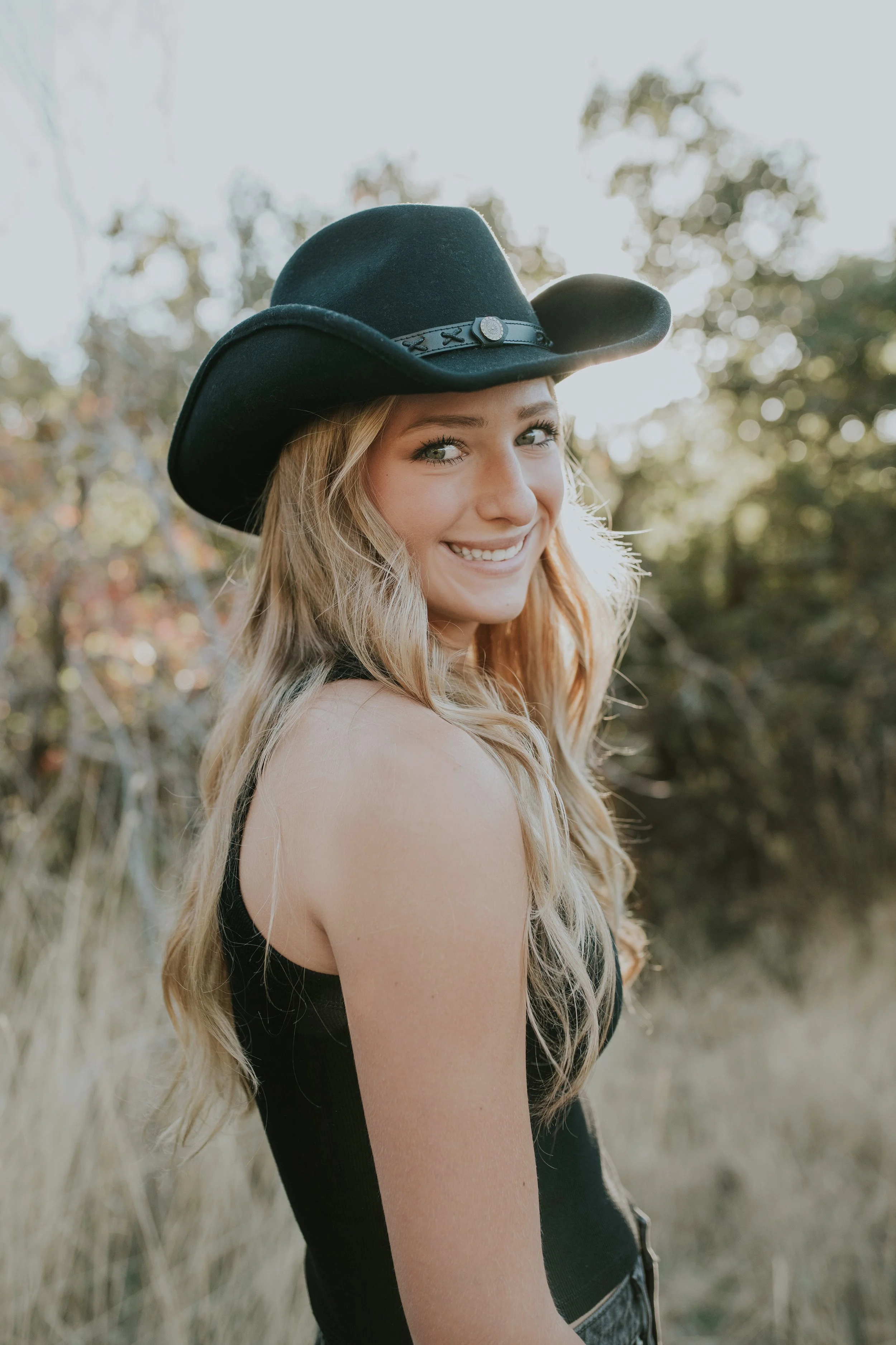 A smiling young woman with long blonde hair wearing a black cowboy hat and black sleeveless top outdoors with blurred trees in the background.