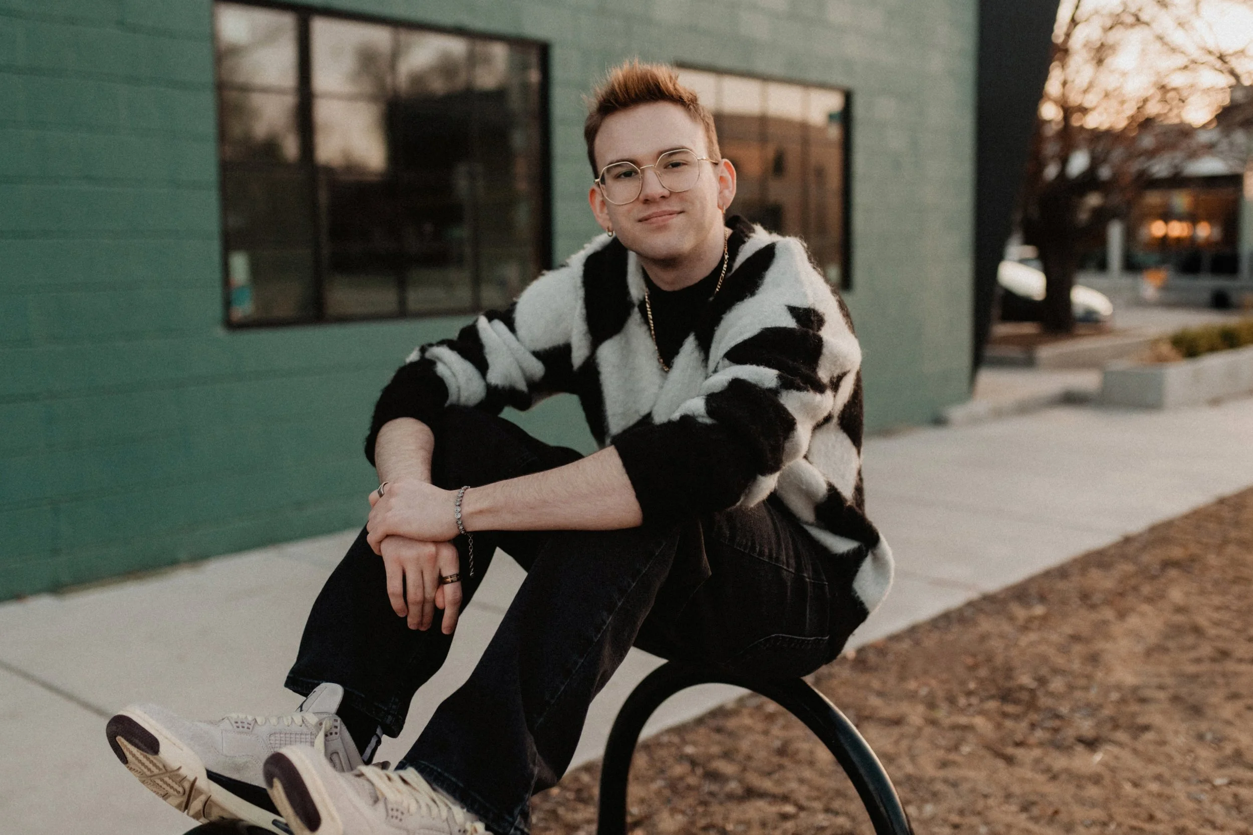 A young man with glasses and earrings sitting on a bike outside near a green building, with trees and cars in the background, during sunset.