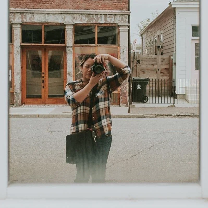 Woman taking a photo of her reflection in a mirror outdoors, with buildings and a street in the background.