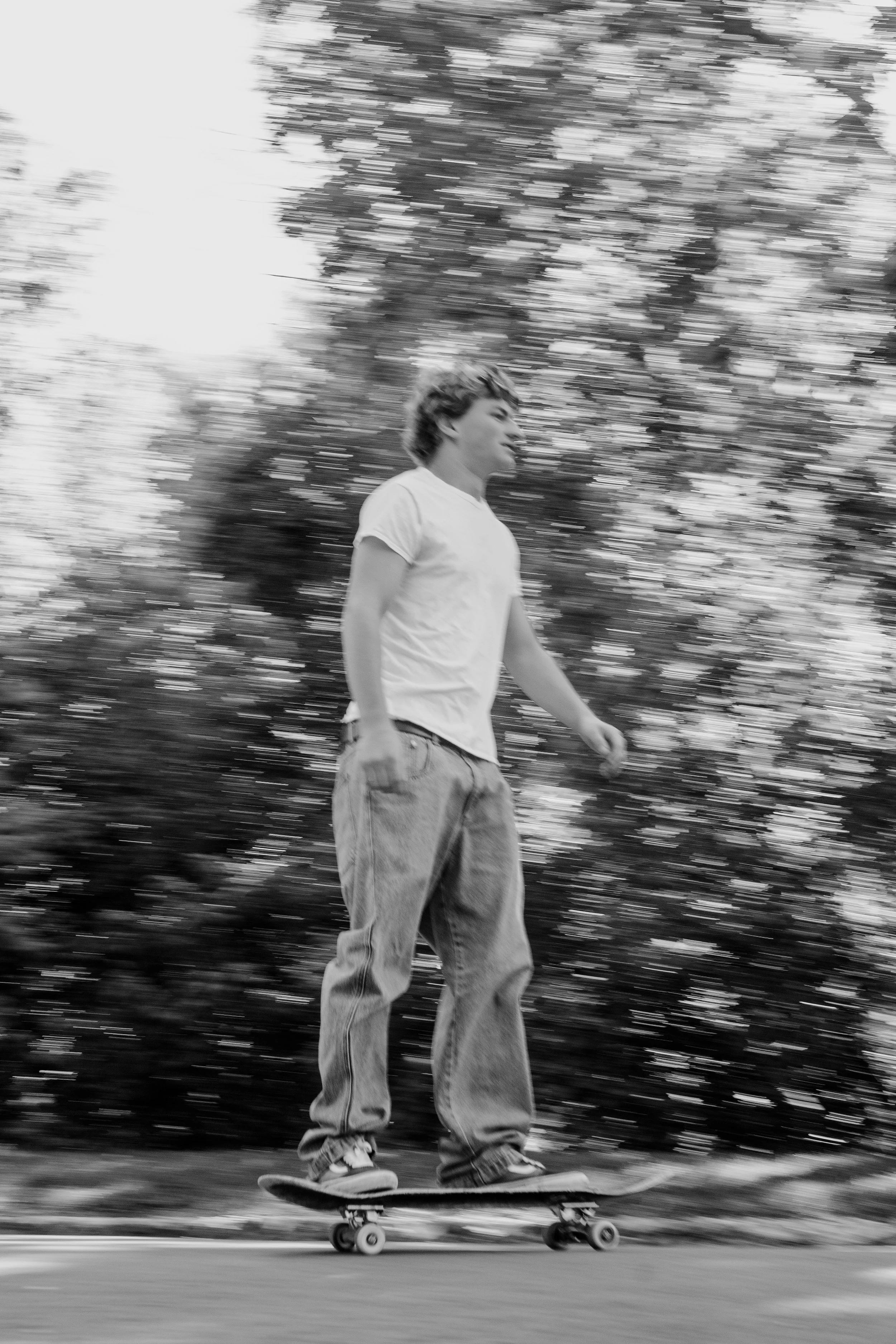 A young person skateboarding outdoors during daytime, with trees and sky in the background, captured in black and white with motion blur.