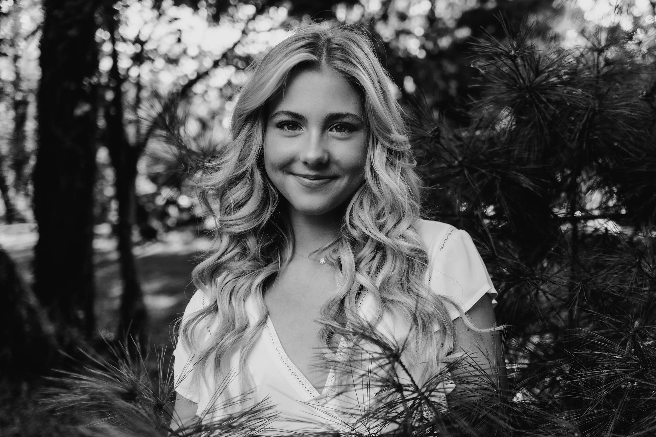 A young woman with long curly hair smiling outdoors, surrounded by trees and foliage in black and white.