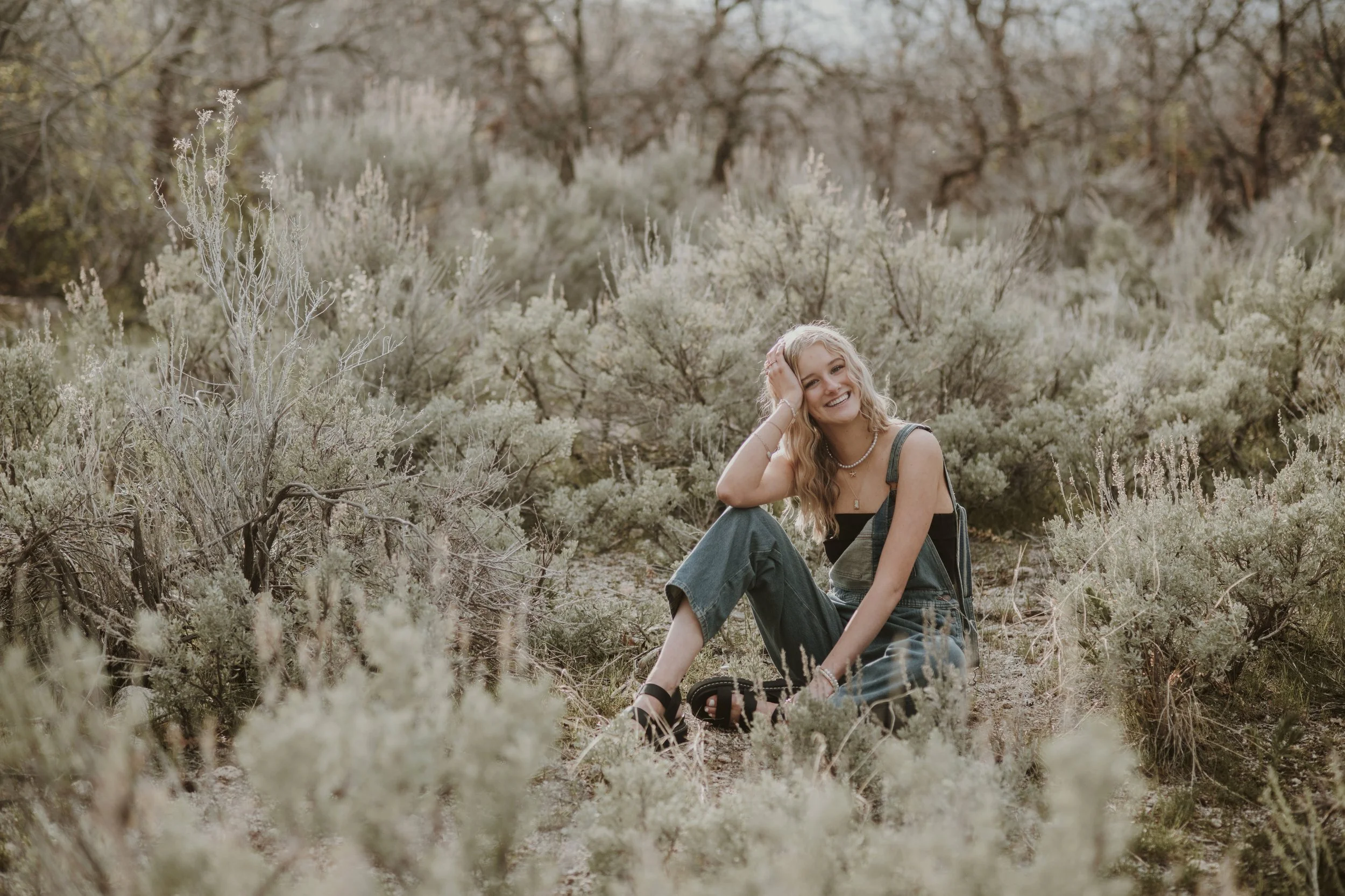 A young woman with blonde hair, sitting on the ground among dry shrubs, smiling and wearing a black top, green overalls, and sandals in a natural outdoor setting.
