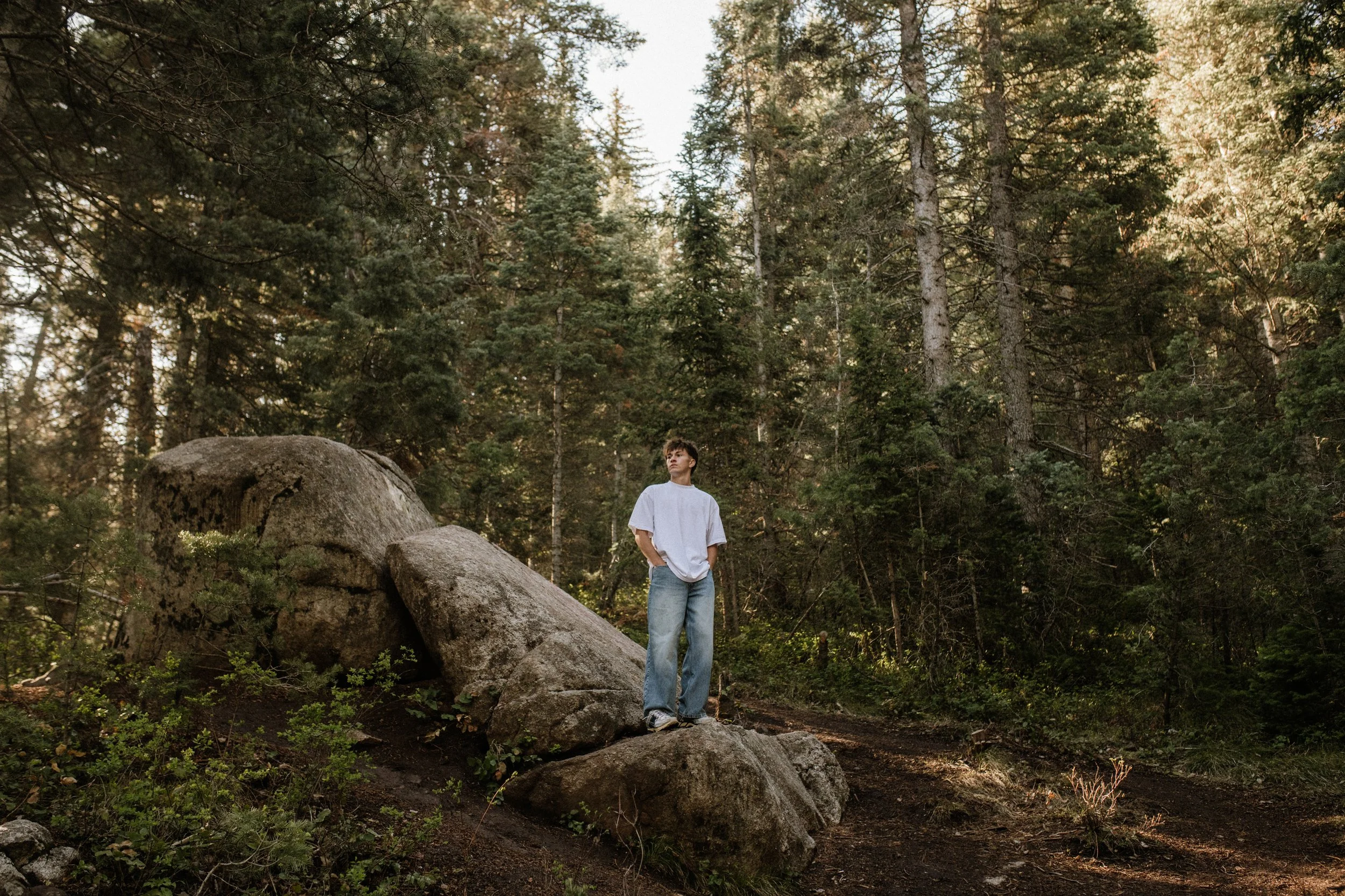A young man with brown hair wearing a loose white t-shirt and blue jeans stands on a large rock in a forested area with tall trees and sunlight filtering through the branches.