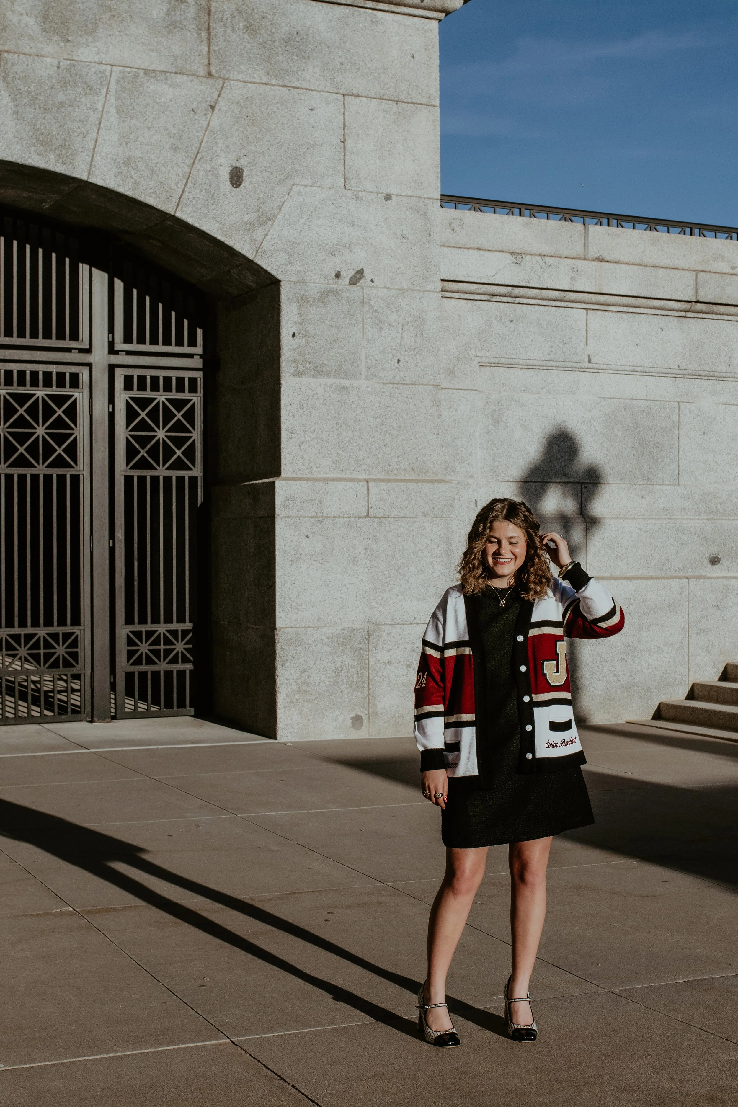 A young woman with curly hair smiling, wearing a varsity jacket, black dress, and high heels, standing outside in shadow on a sunny day in front of a large stone building with stairs and railing.