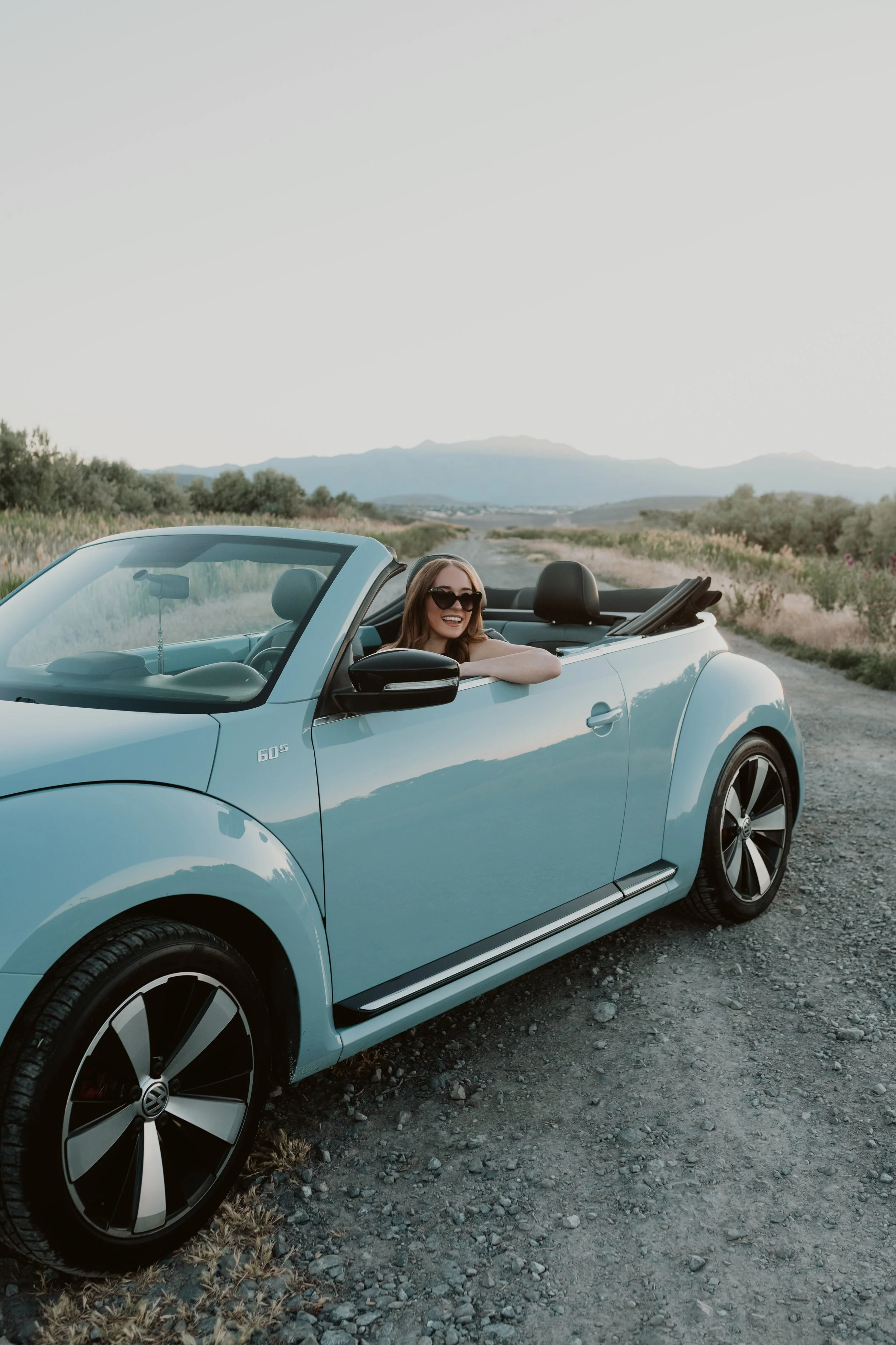 A woman with long hair and sunglasses sitting in the driver's seat of a light blue convertible car, smiling, in a rural landscape with mountains in the background.