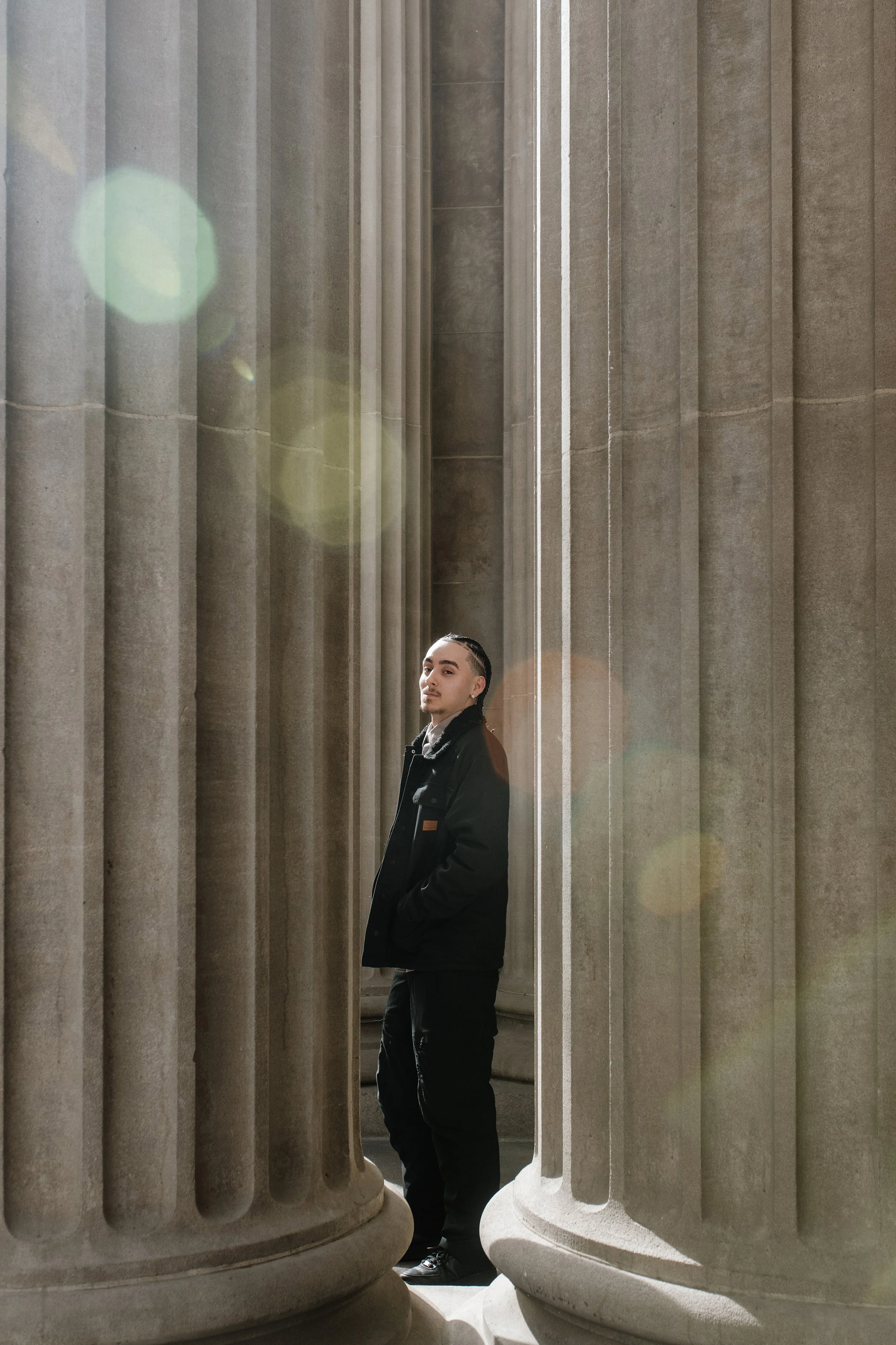 A young man with short dark hair standing between large stone columns, wearing a black jacket, in front of a classical building interior.