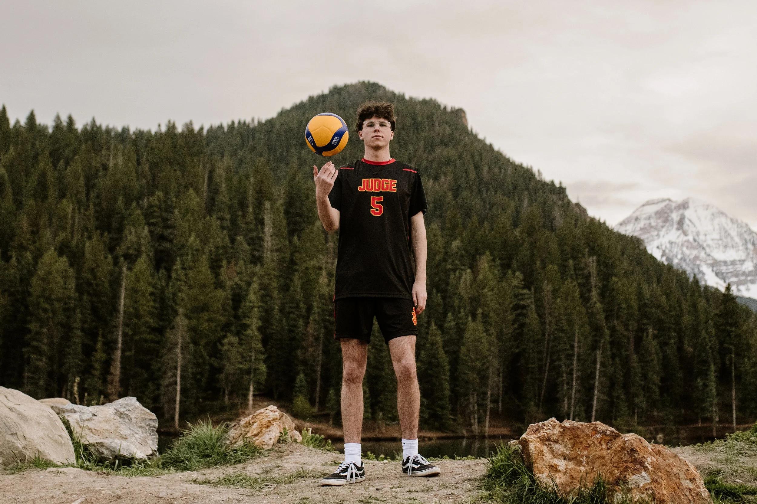 A young man standing outdoors on rocks with a volleyball, mountain and forest background.
